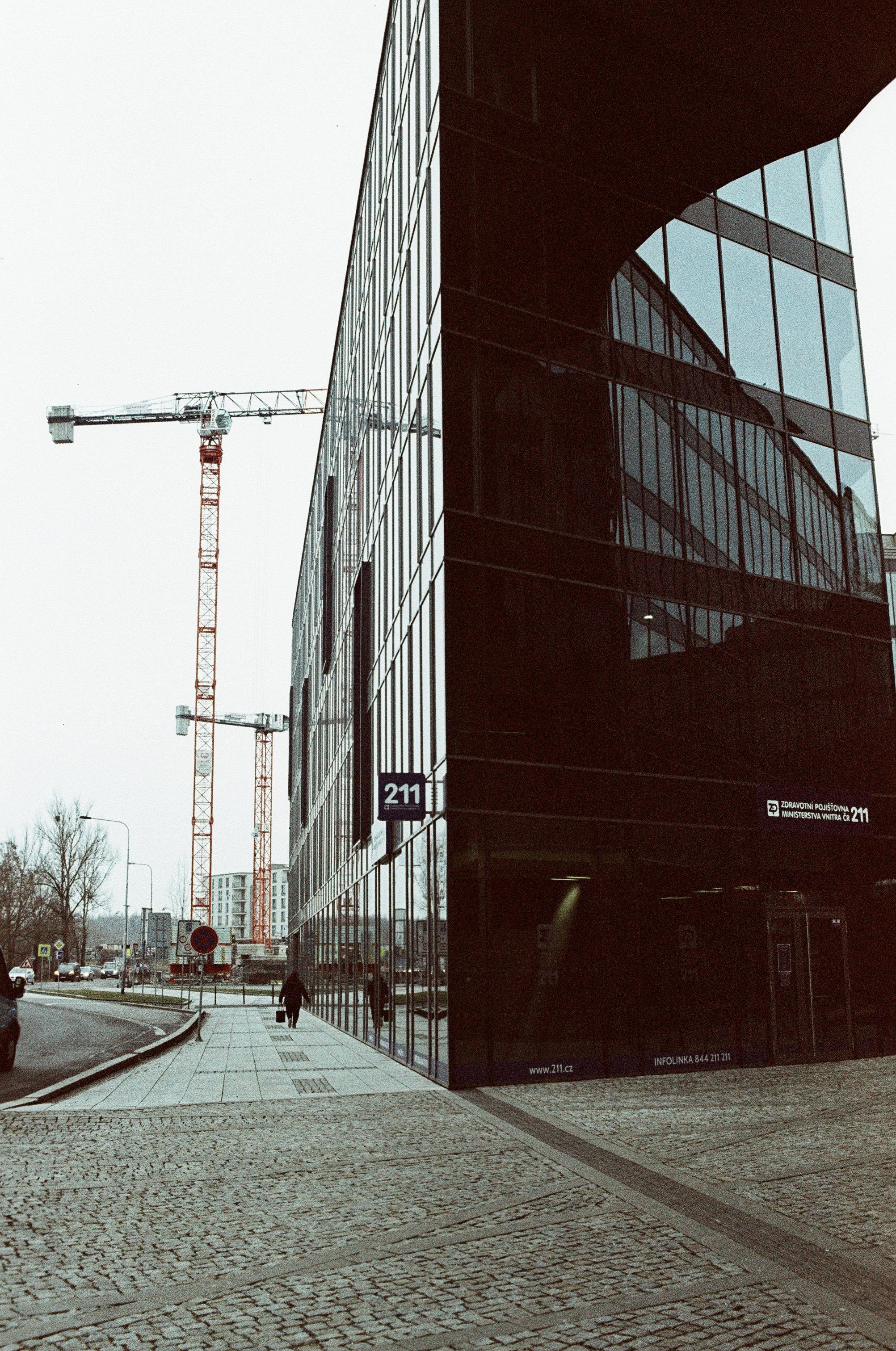 The image shows a modern glass office building with reflective windows. There are two construction cranes visible in the background, indicating ongoing construction in the area. The foreground features a cobblestone sidewalk and a person walking. The building is located on street number 211, as indicated by the signage.
