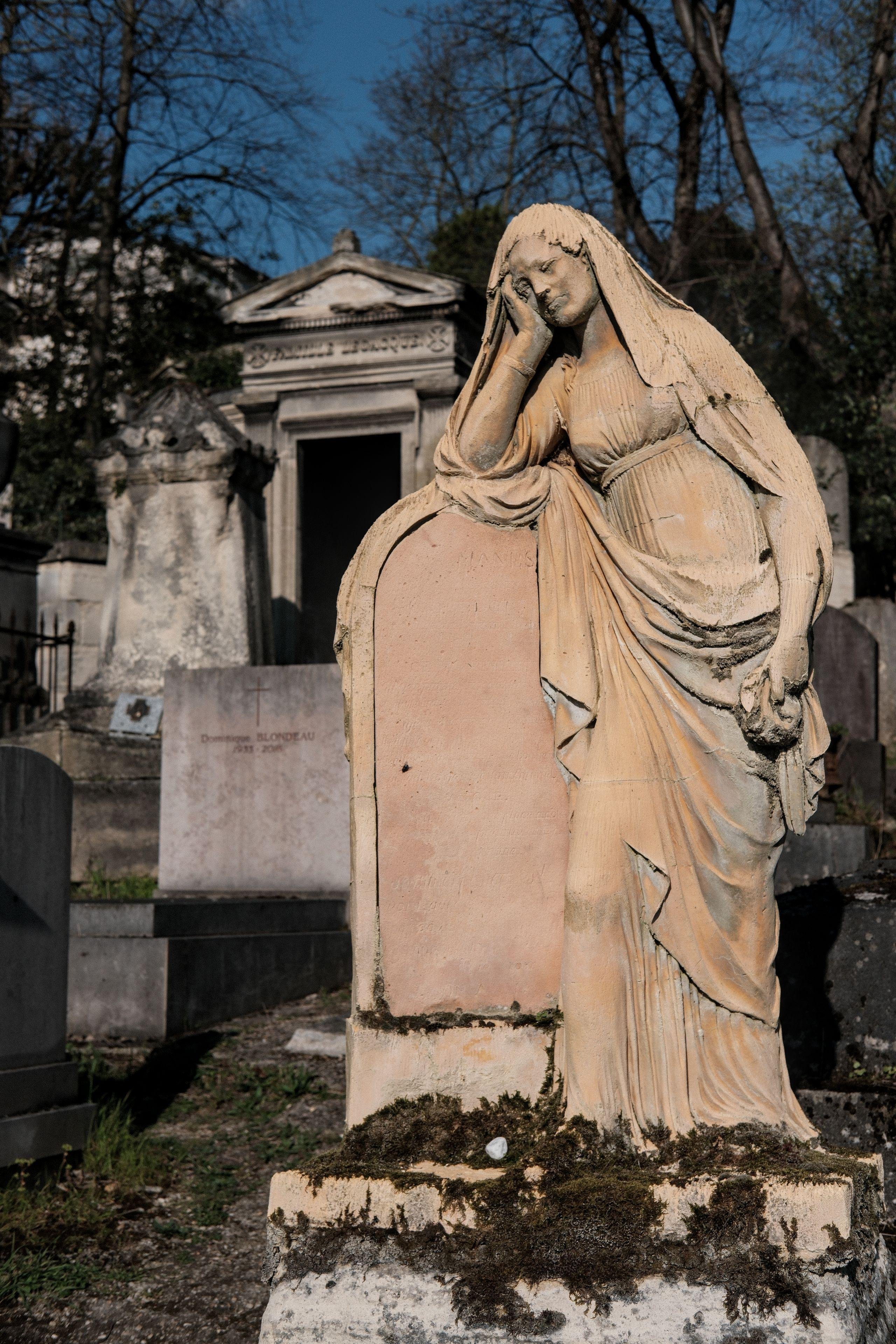 Photo verticale. Au premier plan, une statue de femme en toge et avec un voile posé sur la tête, appuyant son coude droit sur une stèle sans inscription. Derrière, des tombes et des mausolées sous un ciel bleu