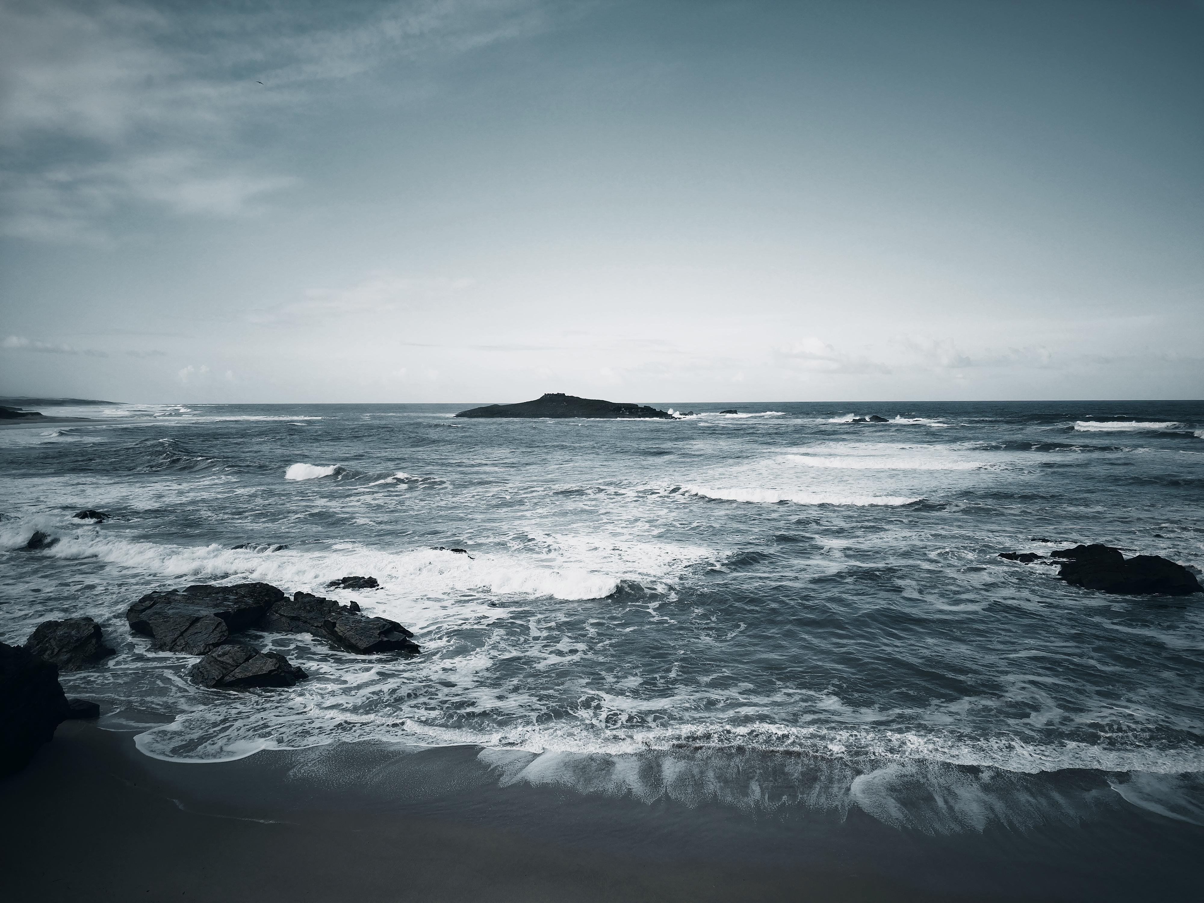 This image depicts a seascape with a rocky shoreline in the foreground. Waves are crashing onto the rocks, and there's an island or large rock formation in the distance. The scene has a calm, overcast atmosphere.