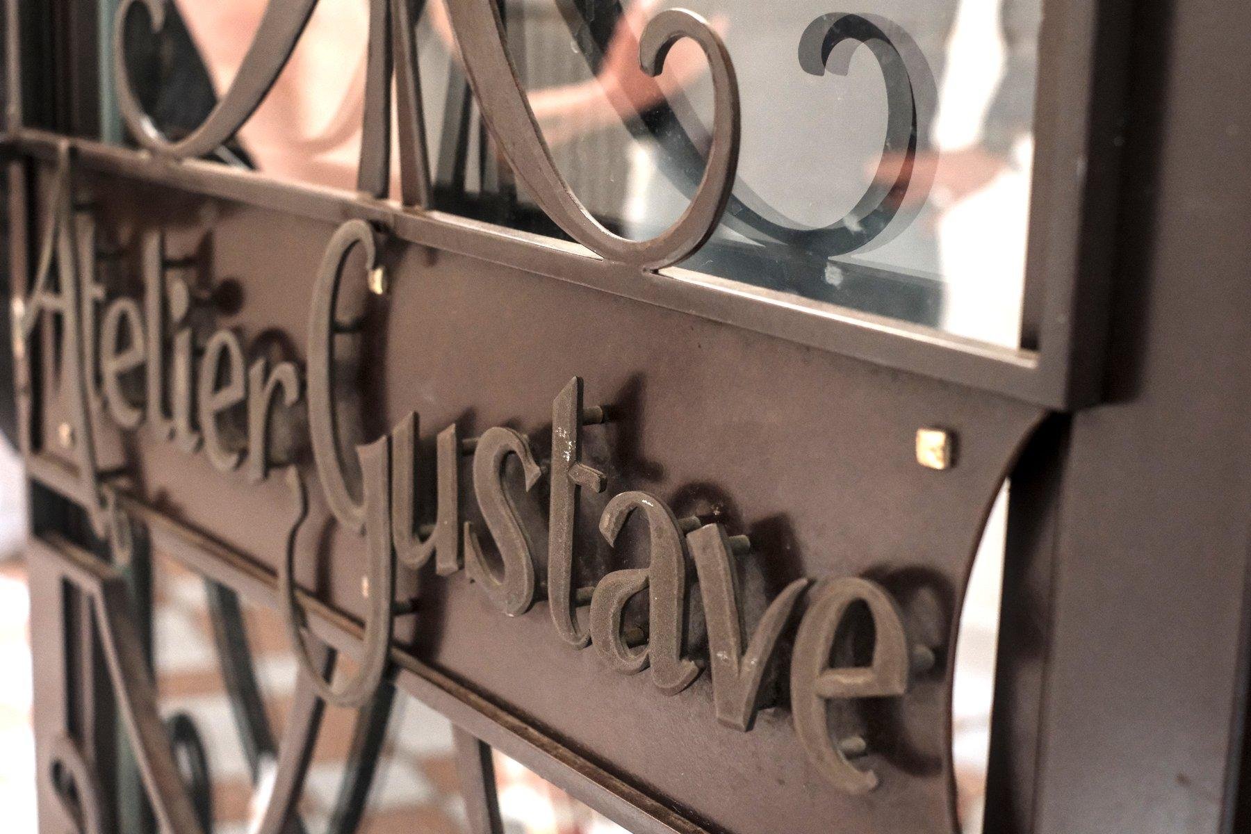 Photo horizontale. Gros plan sur la décoration en fer forgé marron d’une porte, avec les lettres Atelier Gustave en typo artistique
