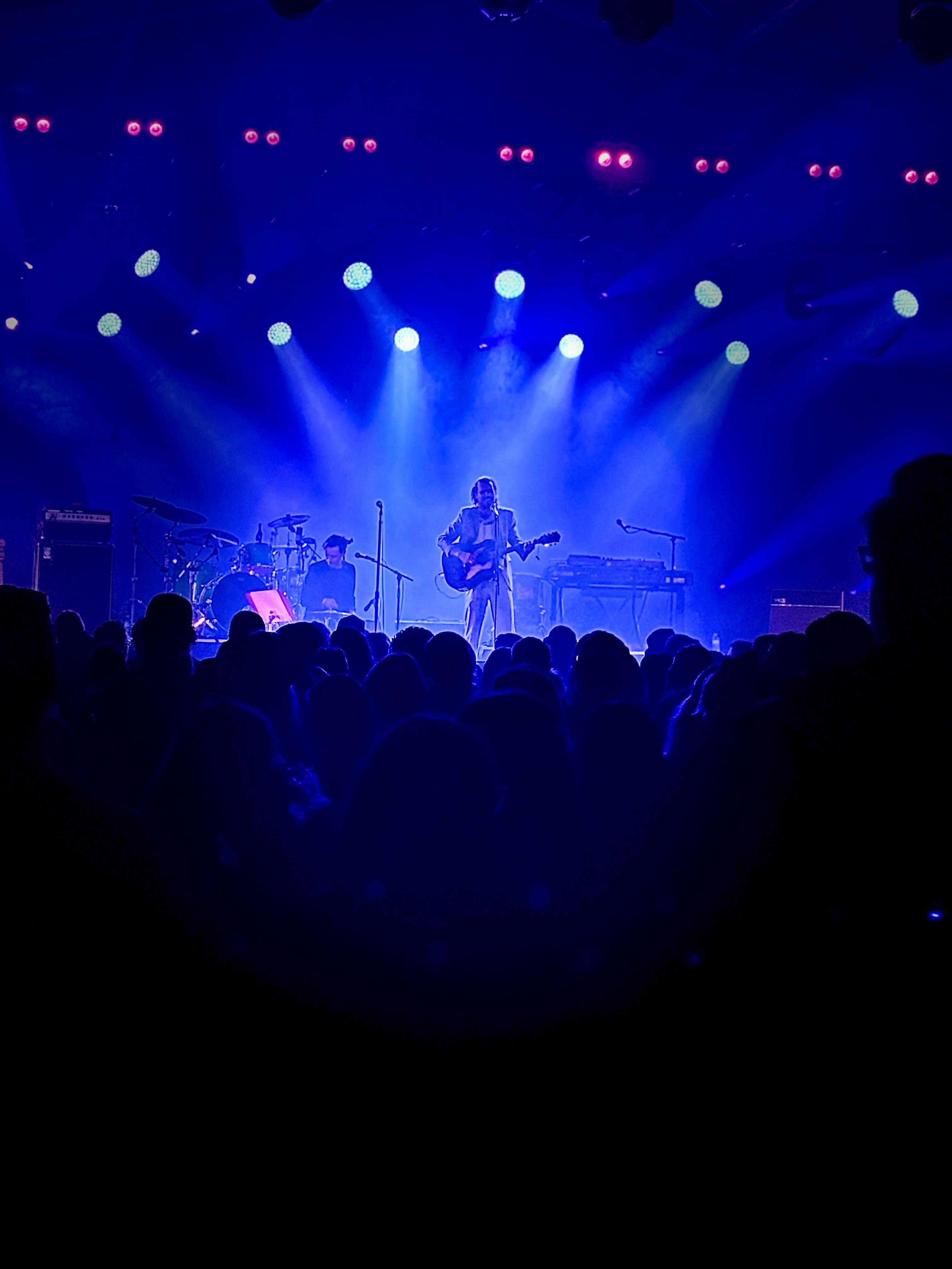 Concert stage lit with blue spotlights; a guitarist stands at center microphone, drummer and drum kit on the left, keyboard setup on the right, and a silhouetted audience in the foreground.