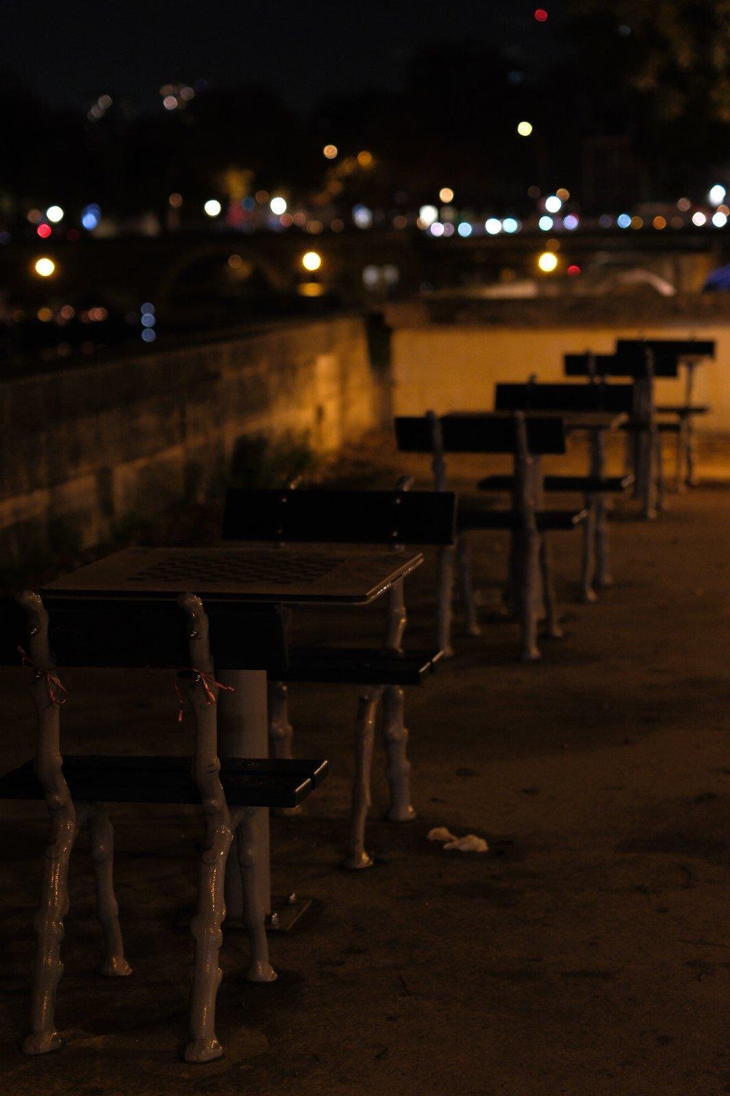 Photo verticale de nuit. Une rangée de tables extérieure à côté d’une rambarde en pierre qui ont toutes 2 chaises face à face et une plateau d’échec intégré. La scène est éclairée par une lumière orange et des points lumineux flou sont en arrière plan