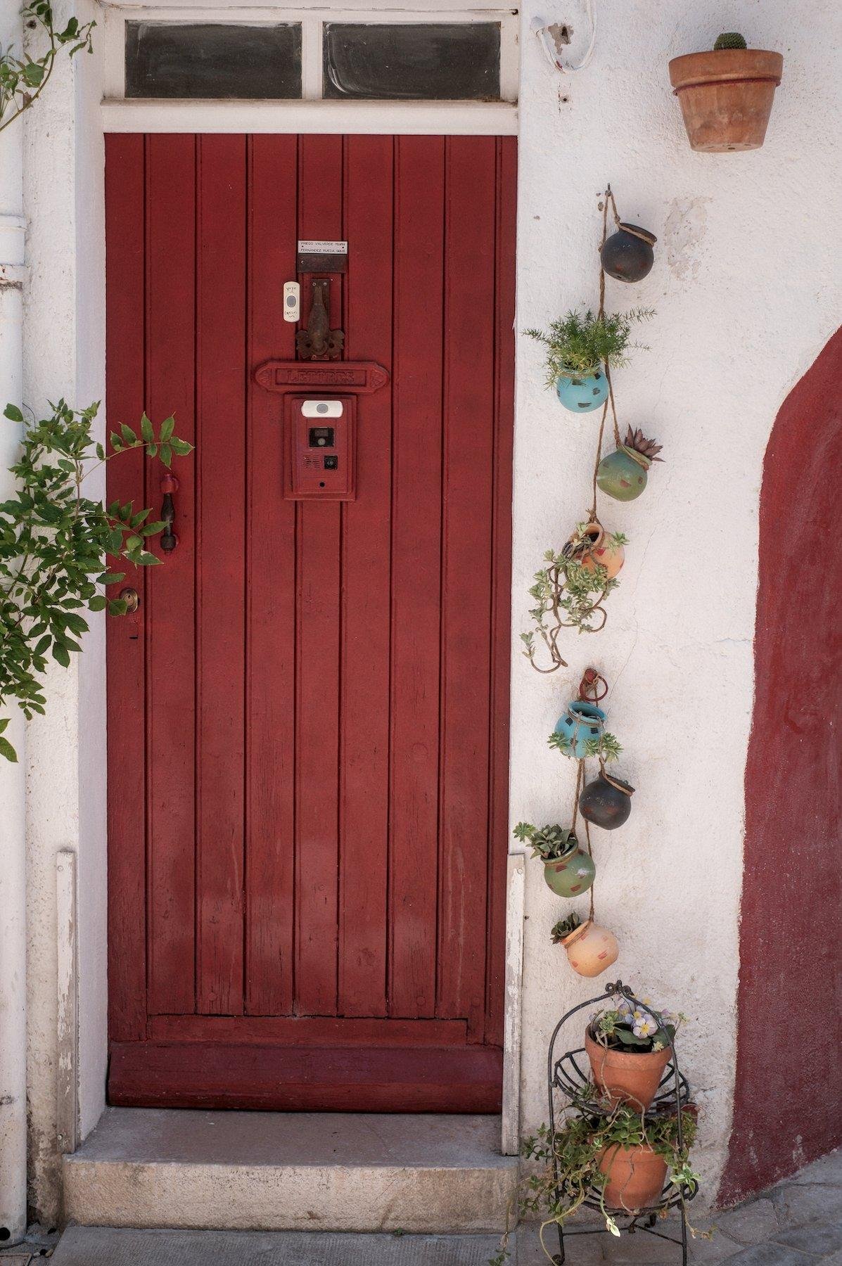 Photo verticale. Une porte rouge en bois avec à la place du judas, un système de caméra. Sur le mur blanc à droite, une guirelande verticale de petits pots de fleurs avec des plantes grasses dedans