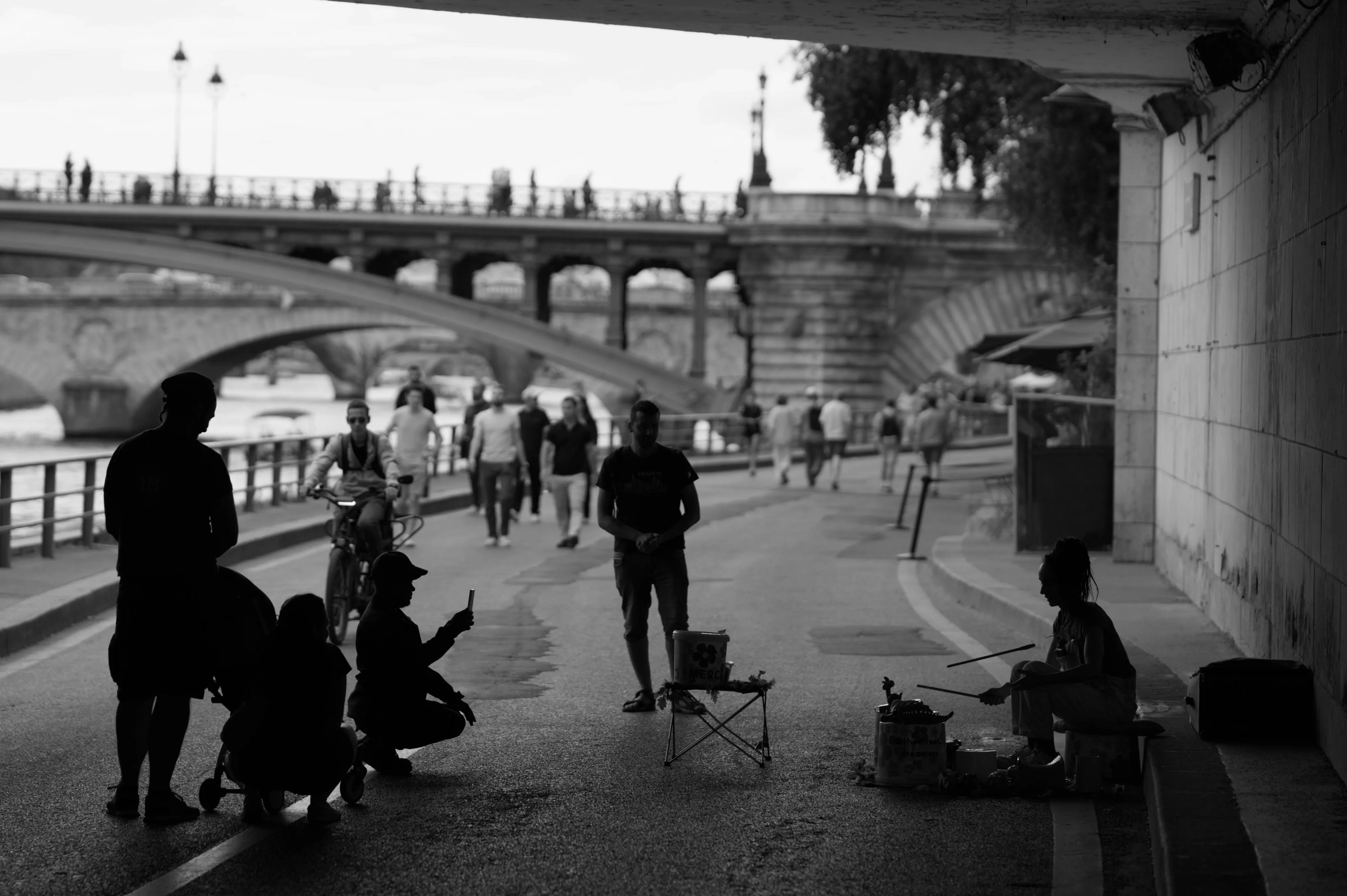 Monochrome black-and-white photograph of passerbys watching a street percussionist on a riverside promenade. The musician and the handful of people who are her audience have only the shape of their bodies visible. Their features are hidden by backlight, as they are under a bridge and so is the camera. In the background, out of focus, more passerbys are walking on the promenade and the river, as well as other stone bridges, can be seen further away.