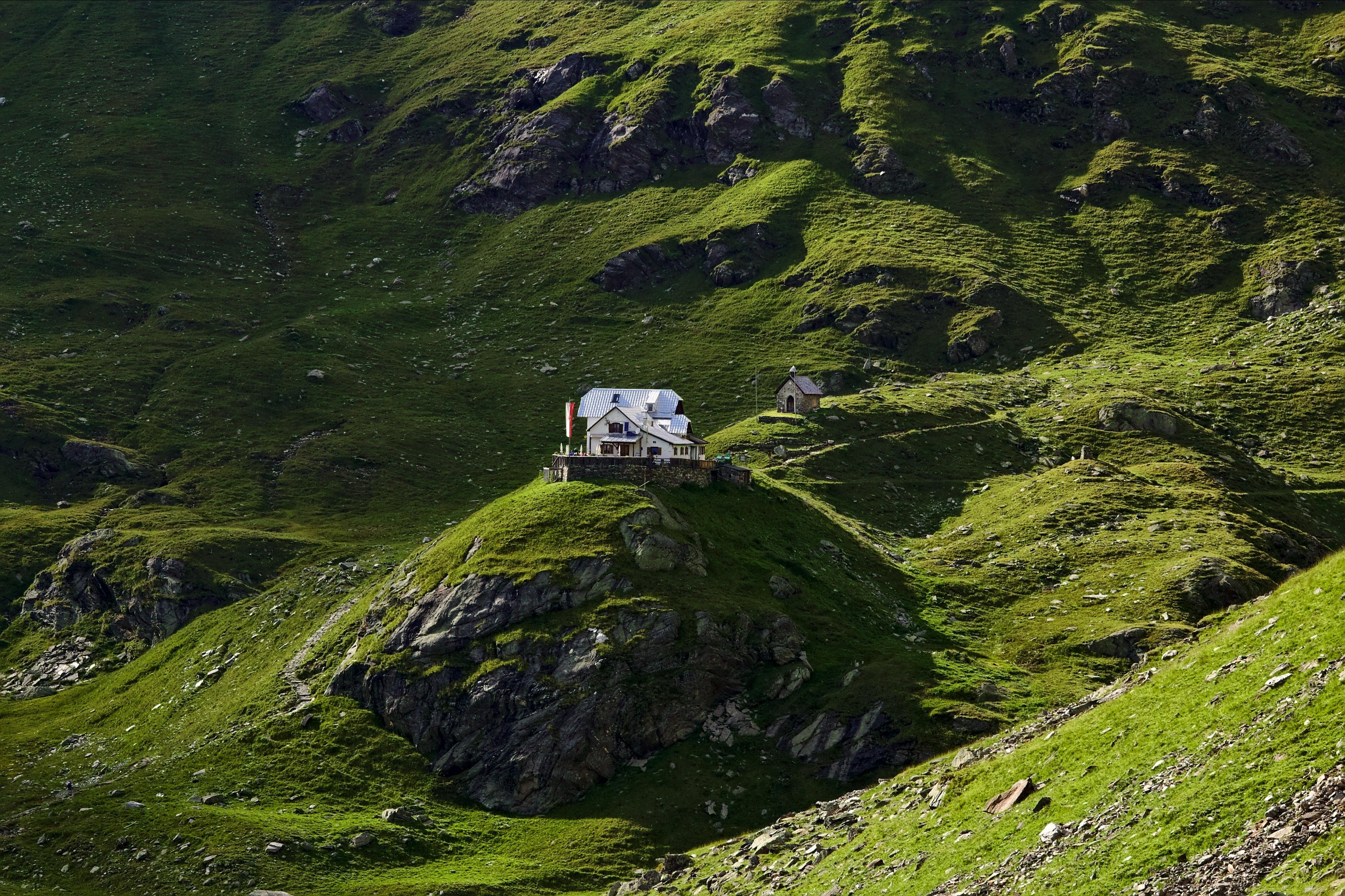 A white mountain lodge exposedly standing on a grassy hill or ridge, surrounded by steep rocky and green hillsides dotted with exposed rocks and shadows.
