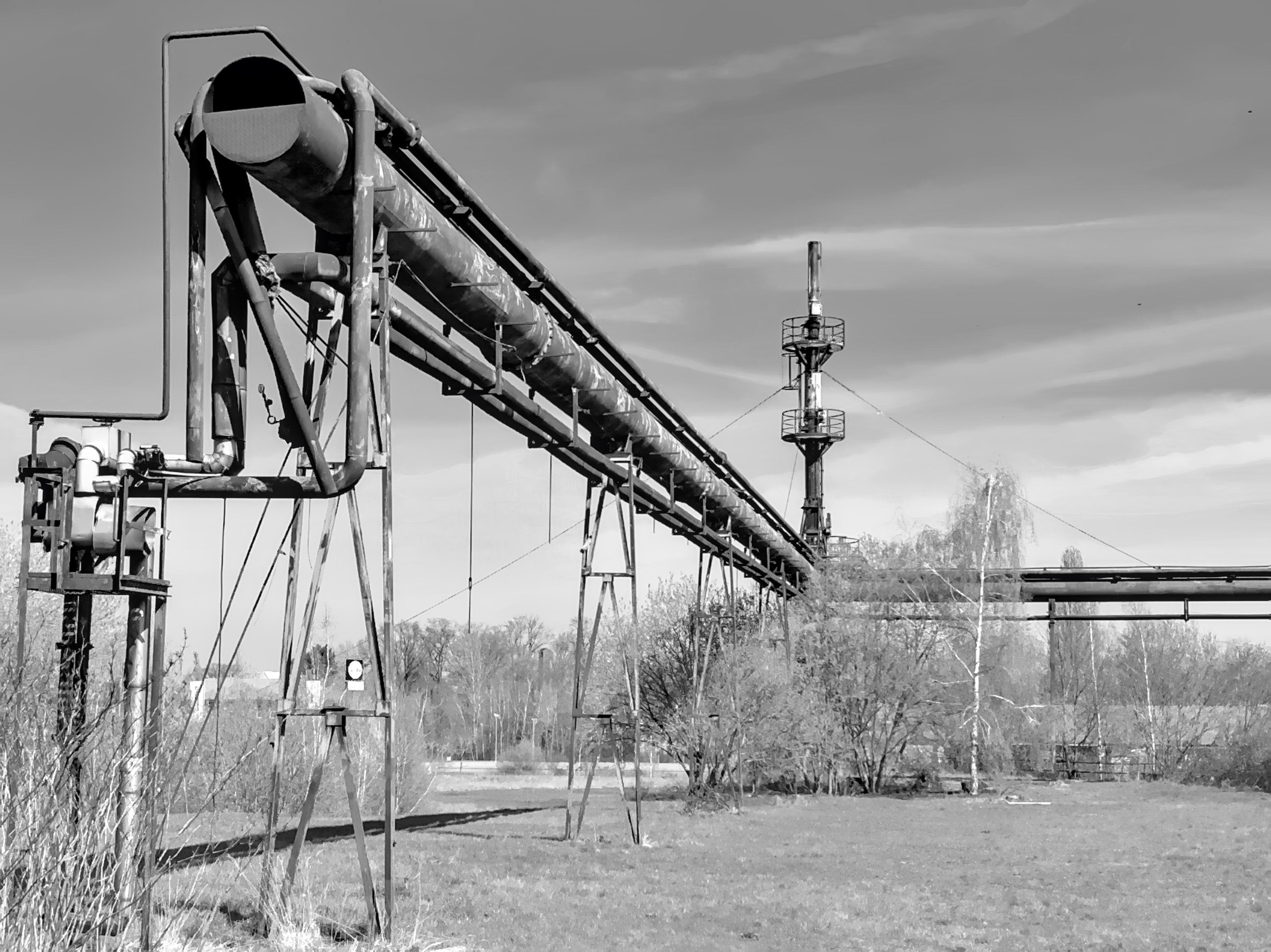 The image depicts a large industrial pipeline structure, elevated and supported by metal frames. It appears to be part of an industrial or processing facility, possibly for transporting fluids or gases. The background includes trees and an open sky, suggesting an outdoor setting.