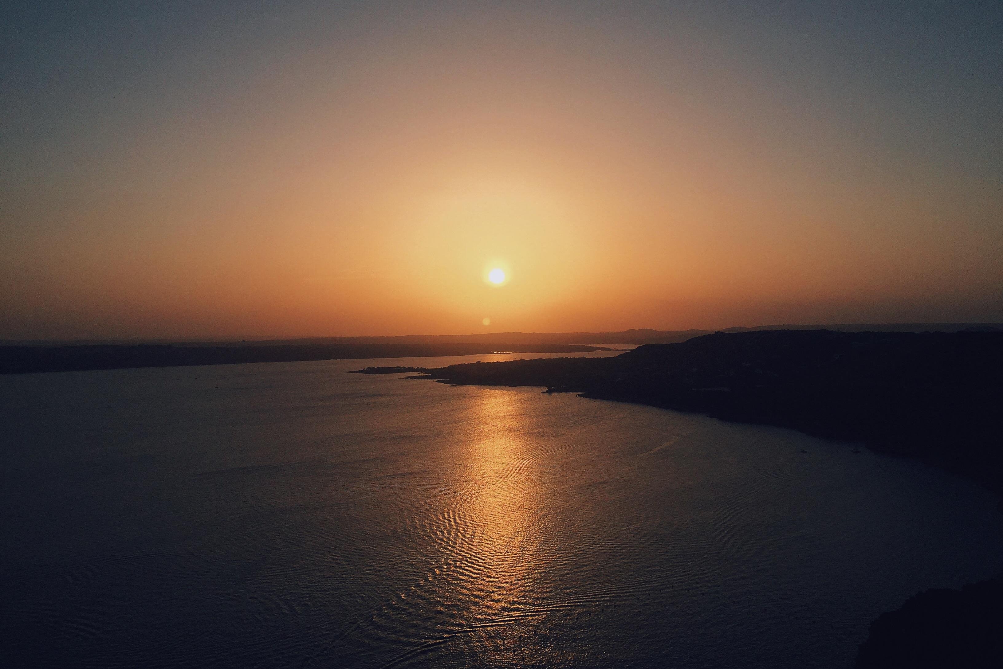 This image shows a sunset over a body of water, with warm colors reflecting on the water's surface. The sun is low on the horizon, creating a serene and peaceful atmosphere. The landscape in the background appears to have some hills or landforms silhouetted against the sky.
