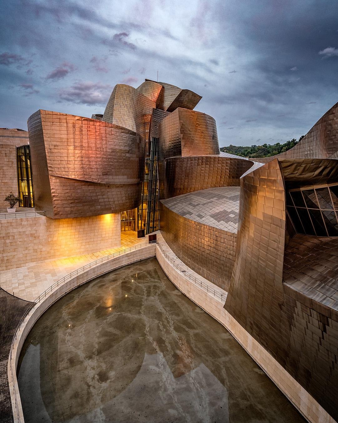 Curved metallic and stone panels form an abstract building façade with a reflective water feature in the foreground. The structure's intricate design features various protruding and overlapping shapes. Evening light casts a warm glow on the coppery surfaces, contrasting with the overcast sky in the background.