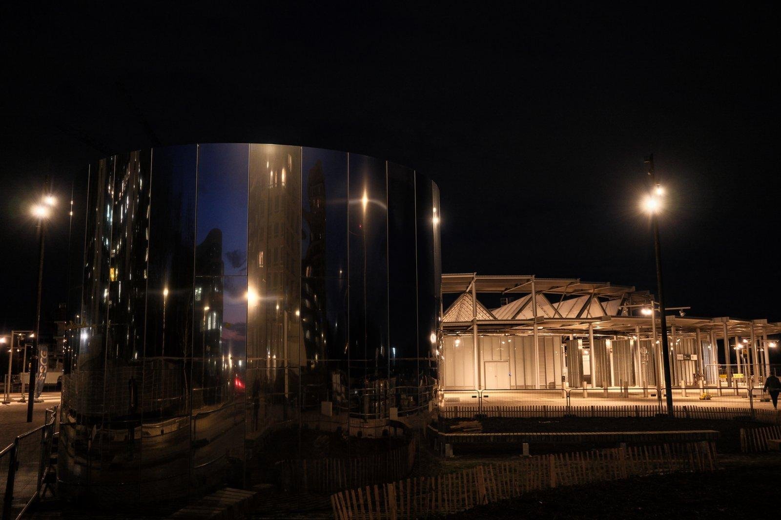 Photo horizontale, de nuit. Au premier plan, une grosse structure cylindrique en acier réfléchissant sur lequel on voit des lampadaires, la route, des gens, tous déformés. Derrière, un bâtiment dont émane beaucoup de lumière orangée