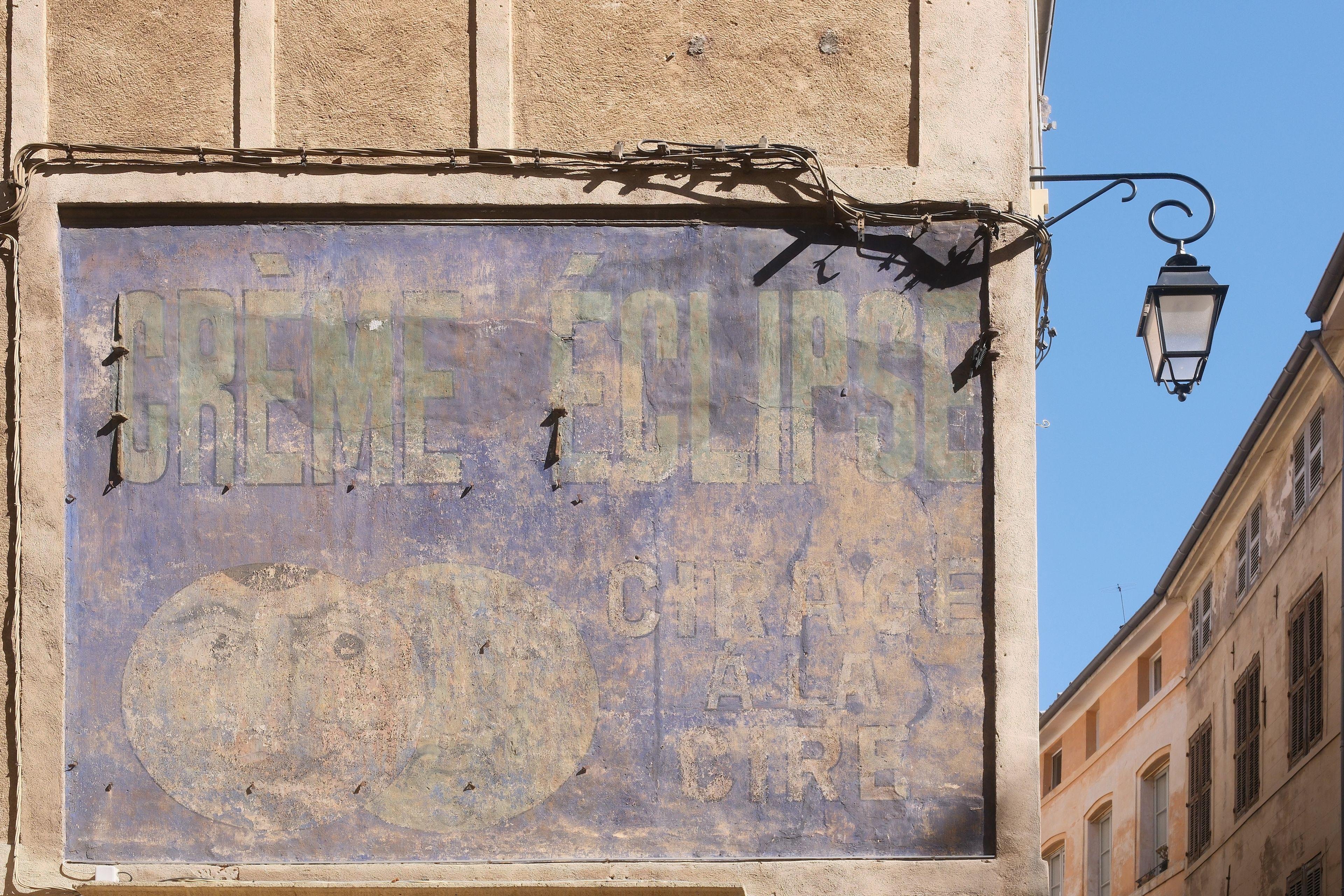 Une vieille publicité peinte sur un immeuble qui a été délavée par le temps. On peut y lire « Crème éclipse. Cirage à la cire ». Sur la droite de cette affiche, un coin de ciel bleu et un lampadaire style vieux lampion à gaz