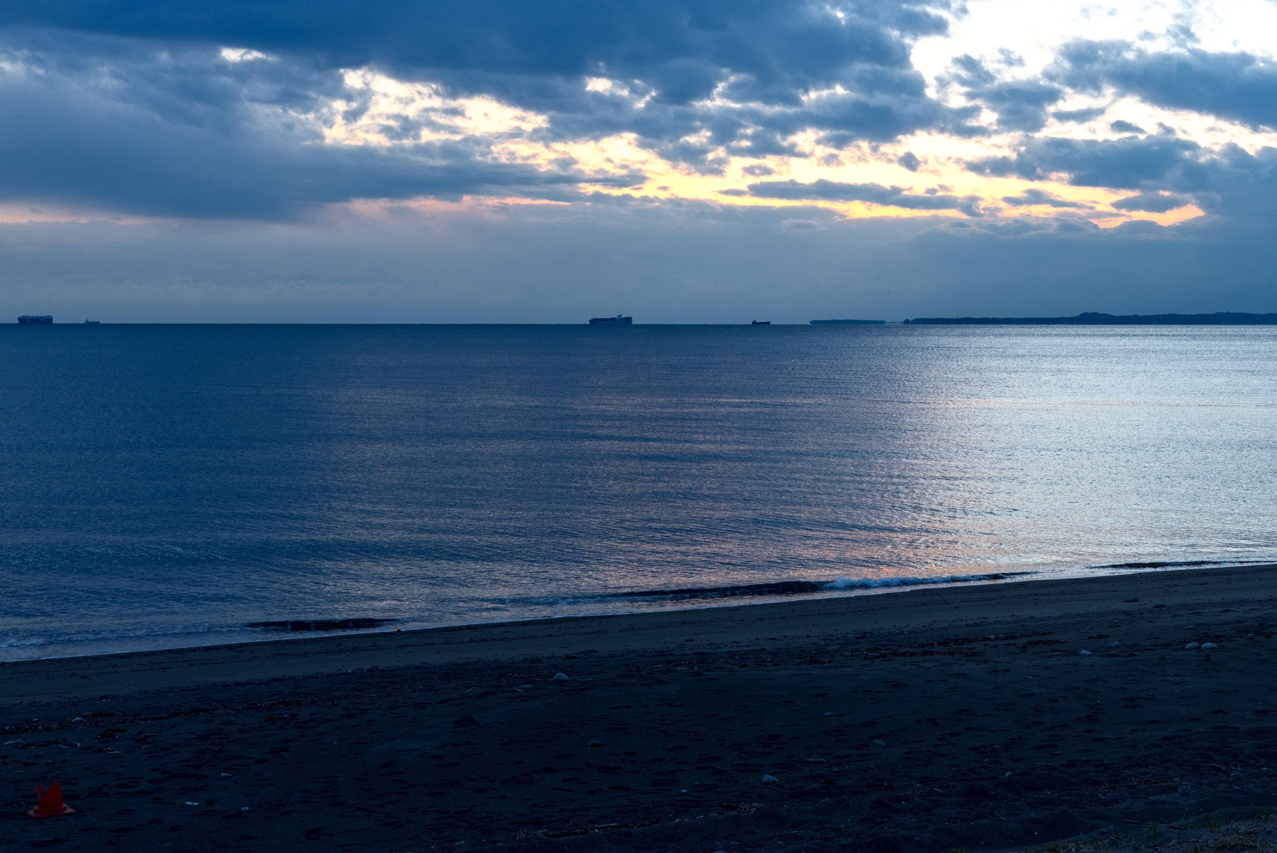 A beach during a cloudy sunset. The dark sand in the forefront heavily contrasts with the lightness of the sky and the bright reflection in the seawater. The clouds have a very cinematic look, interspersed with yellow sunlight. A few ships can be seen in the distance.