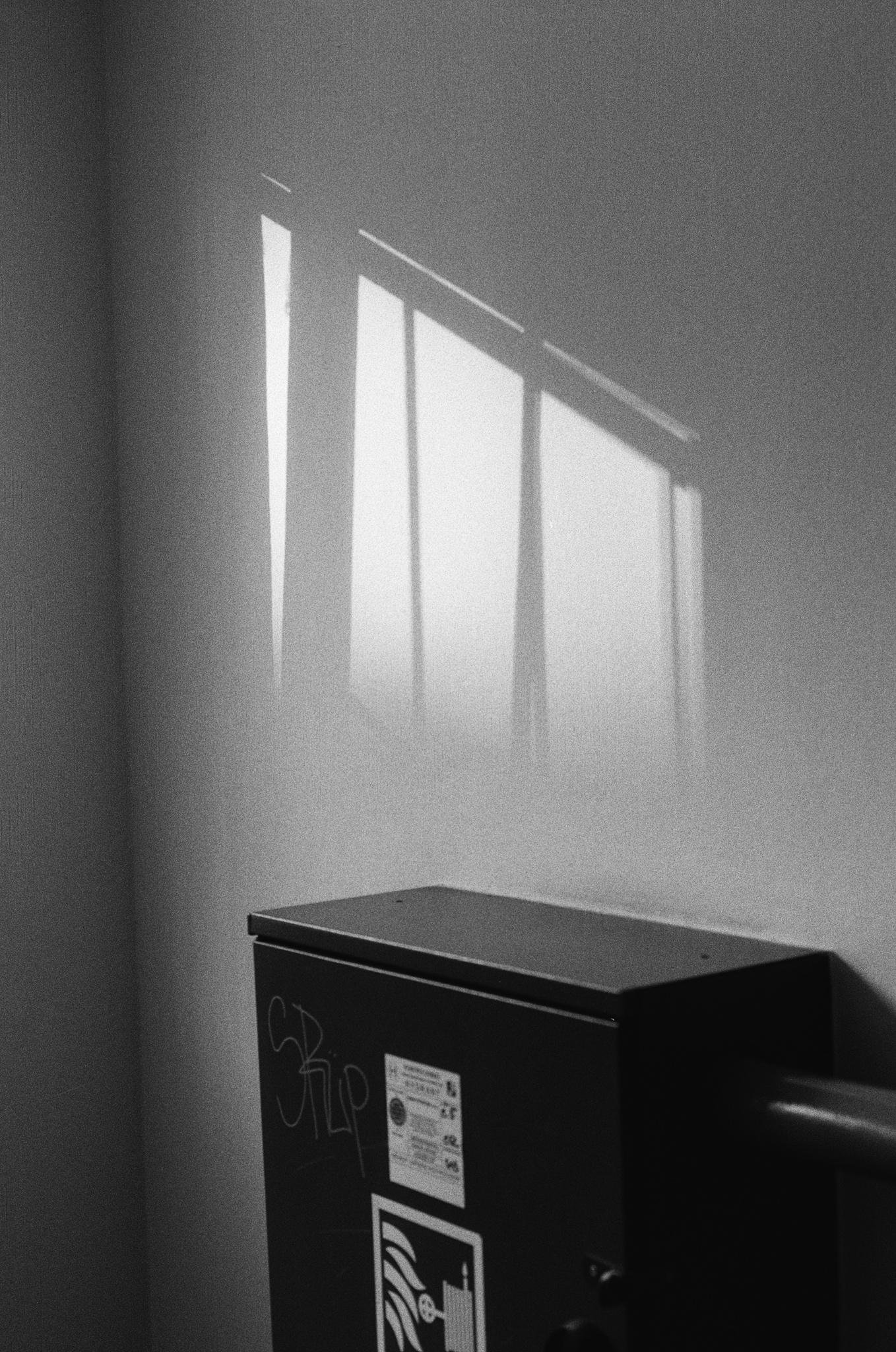 Black-and-white photo of a wall with bright rectangular window shadows cast across it, above a dark metal cabinet or utility box with graffiti and small stickers.