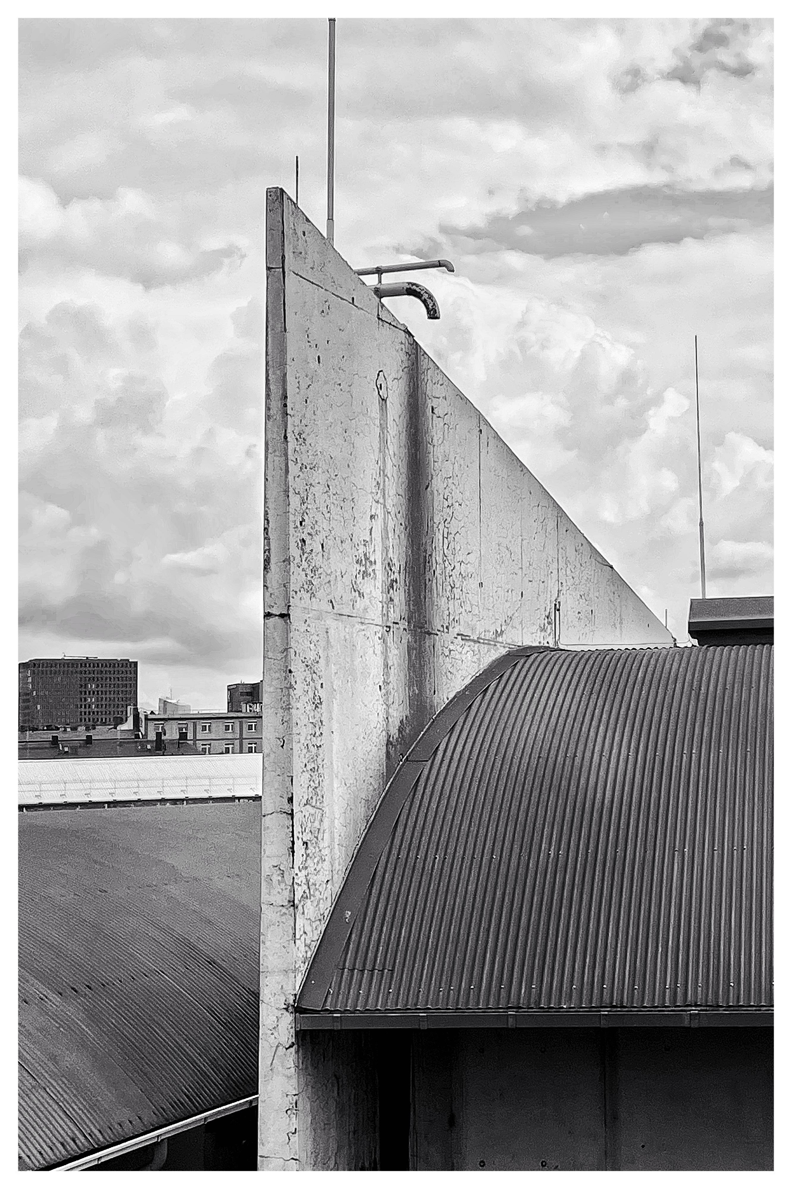 Concrete structure with a triangular, pointed shape and visible cracks, adjacent to a corrugated metal roof. Background includes cloudy sky and distant city buildings. Vertical rods protrude from the top of the structure, with pipes extending over the edge. Black and white color scheme.