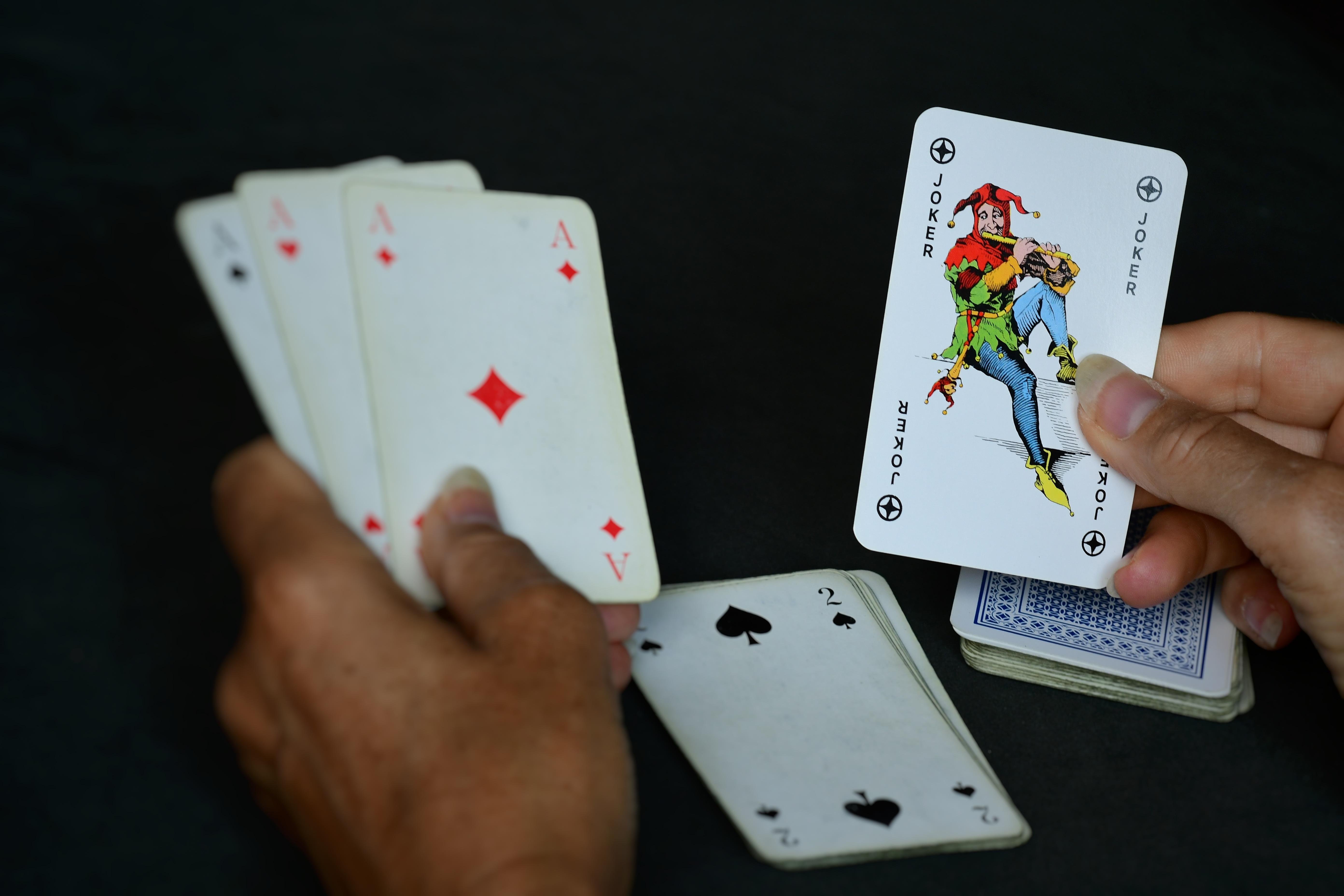 Hands holding a poker hand of five cards, including three aces (clubs, diamonds, hearts) and a two of spades. The right hand holds a joker card with colorful jesters. A deck of cards is partially visible on the table.
###
Hände halten eine Pokerhand mit fünf Karten, darunter drei Asse (Kreuz, Karo, Herz) und eine Pik-Zwei. Die rechte Hand hält eine Jokerkarte mit bunten Narren. Ein Kartenspiel ist teilweise auf dem Tisch sichtbar.