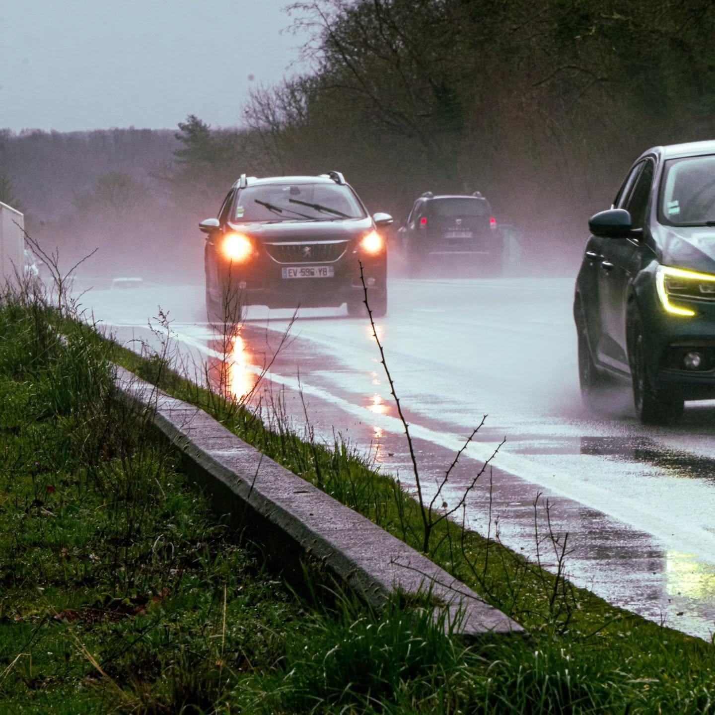 The image shows cars driving on a wet, partially flooded road during a rainy day. The headlights are on, reflecting off the wet surface. There's vegetation on the roadside and trees in the background. The sky is overcast, contributing to the overall misty and damp atmosphere.