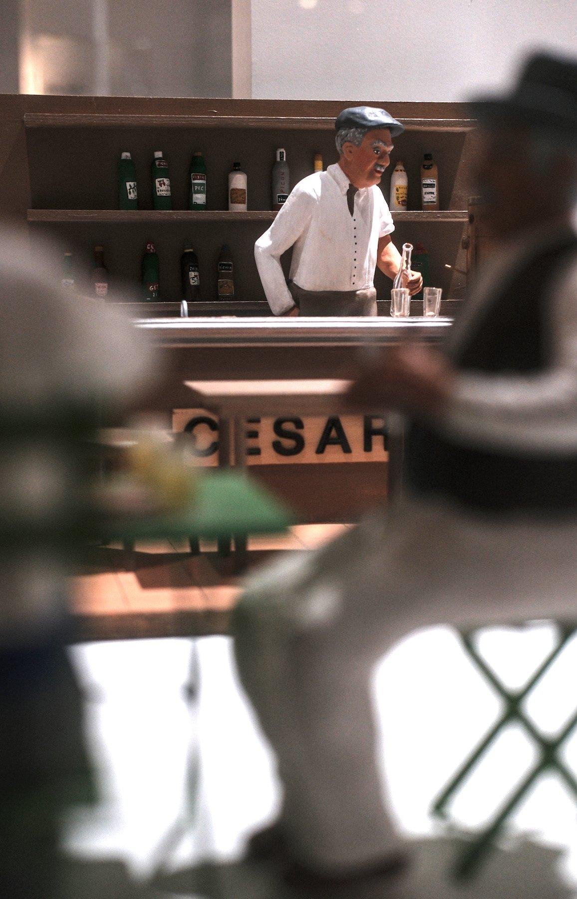Photo verticale. Au premier plan des santons d’hommes assis à une table, floutés, encadrant le sujet de la photo, un santon d’un barman portant un béret, derrière le comptoir