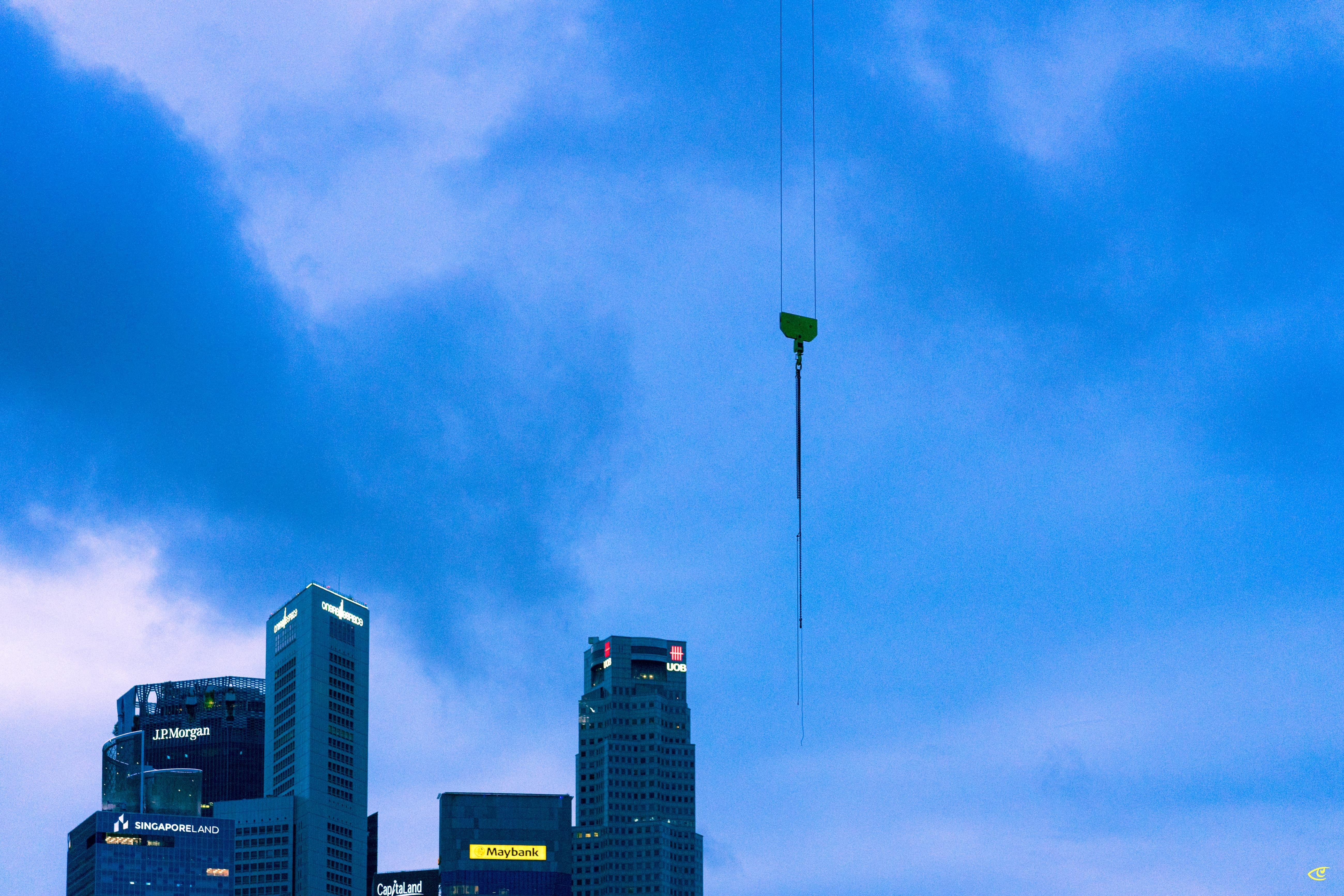 Suspended crane hook hanging from cables against a cloudy blue sky above a city skyline with several high-rise buildings and illuminated signs near the bottom of the frame.