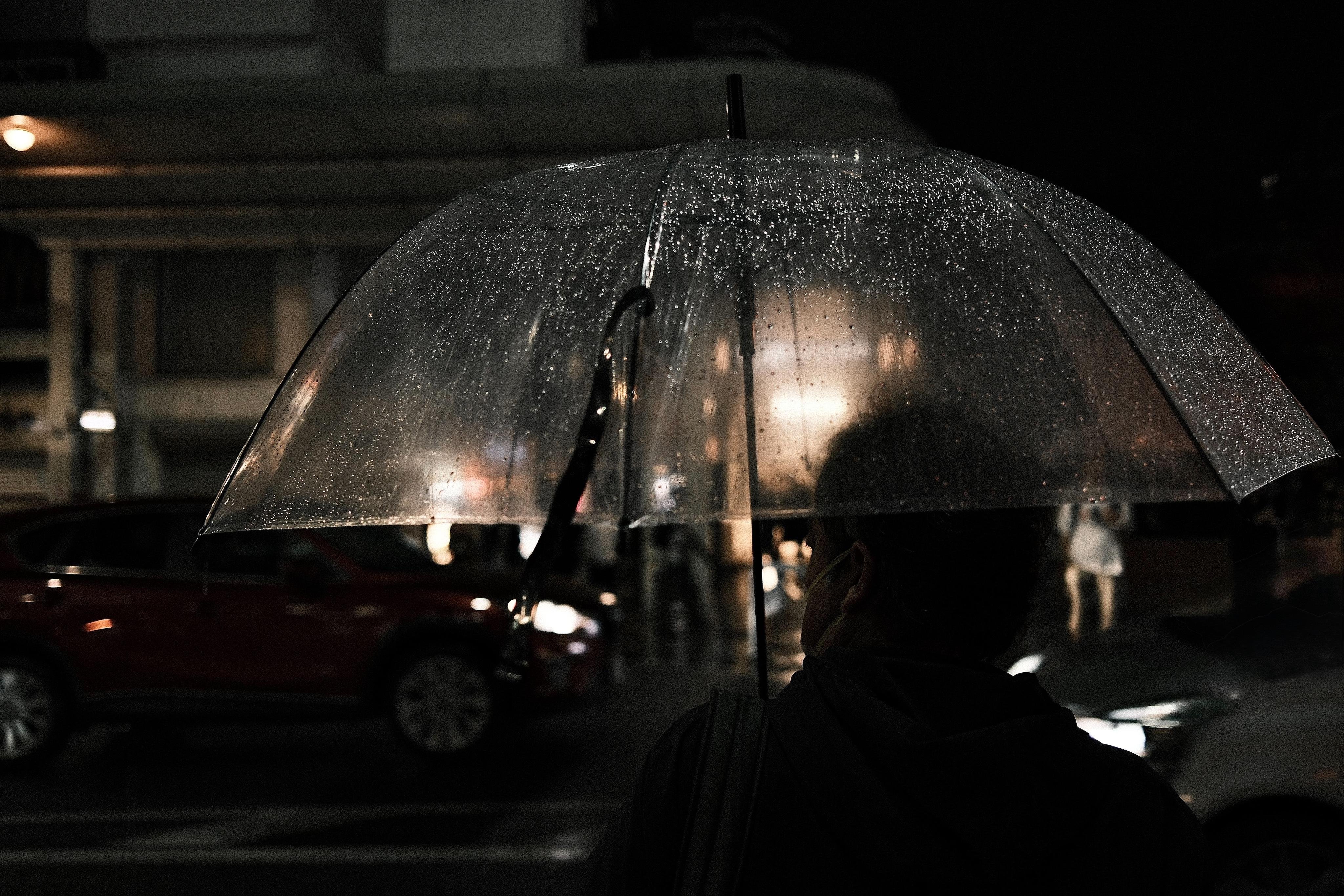 Silhouette of a person holding a clear umbrella dotted with raindrops at night, with blurred streetlights and cars in the background.