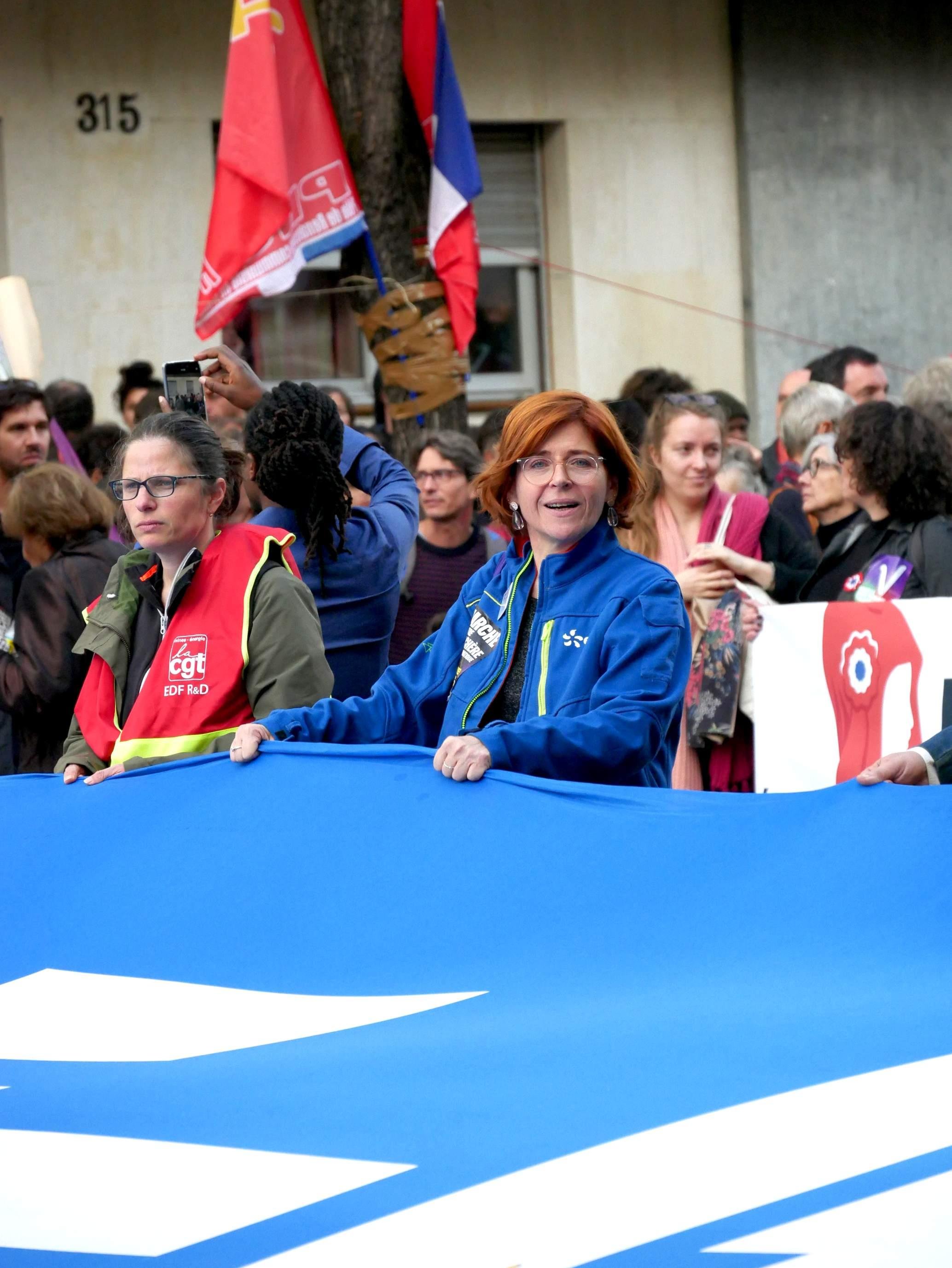 La photo représente un groupe de personnes participant à une manifestation. Ils tiennent une grande bannière bleue et certains participants portent des gilets avec des logos ou des emblèmes. En arrière-plan, il y a différents drapeaux et une foule de personnes. Le cadre semble être urbain, peut-être dans une rue. La personne au centre porte une veste avec le logo EDF sur le torse et regarde l'objectif.