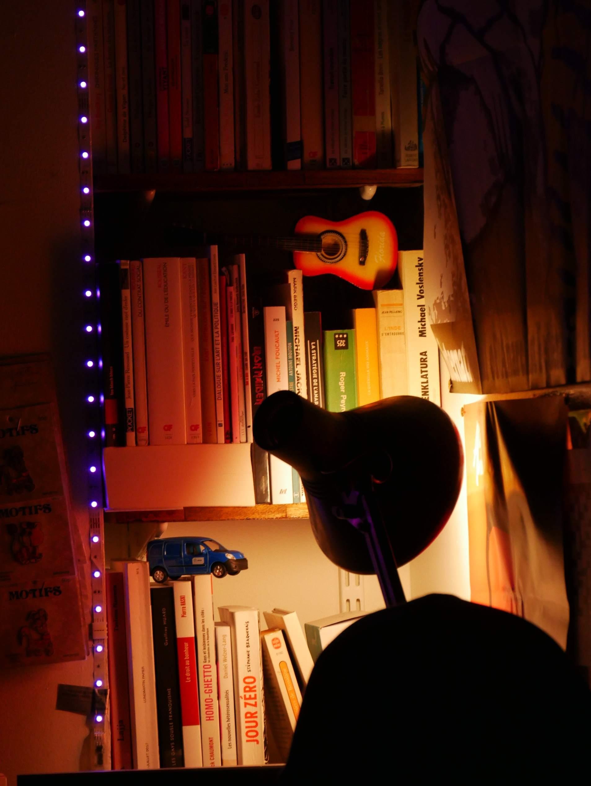 The image features a cozy and warmly-lit bookshelf with various items. There are numerous books arranged on the shelves, and you can see a small model of a guitar as well as a miniature blue car. A desk lamp is positioned in front of the shelf, casting a warm glow on the books. Additionally, there are small purple LED lights running along the side of the shelf, adding a touch of color to the scene.