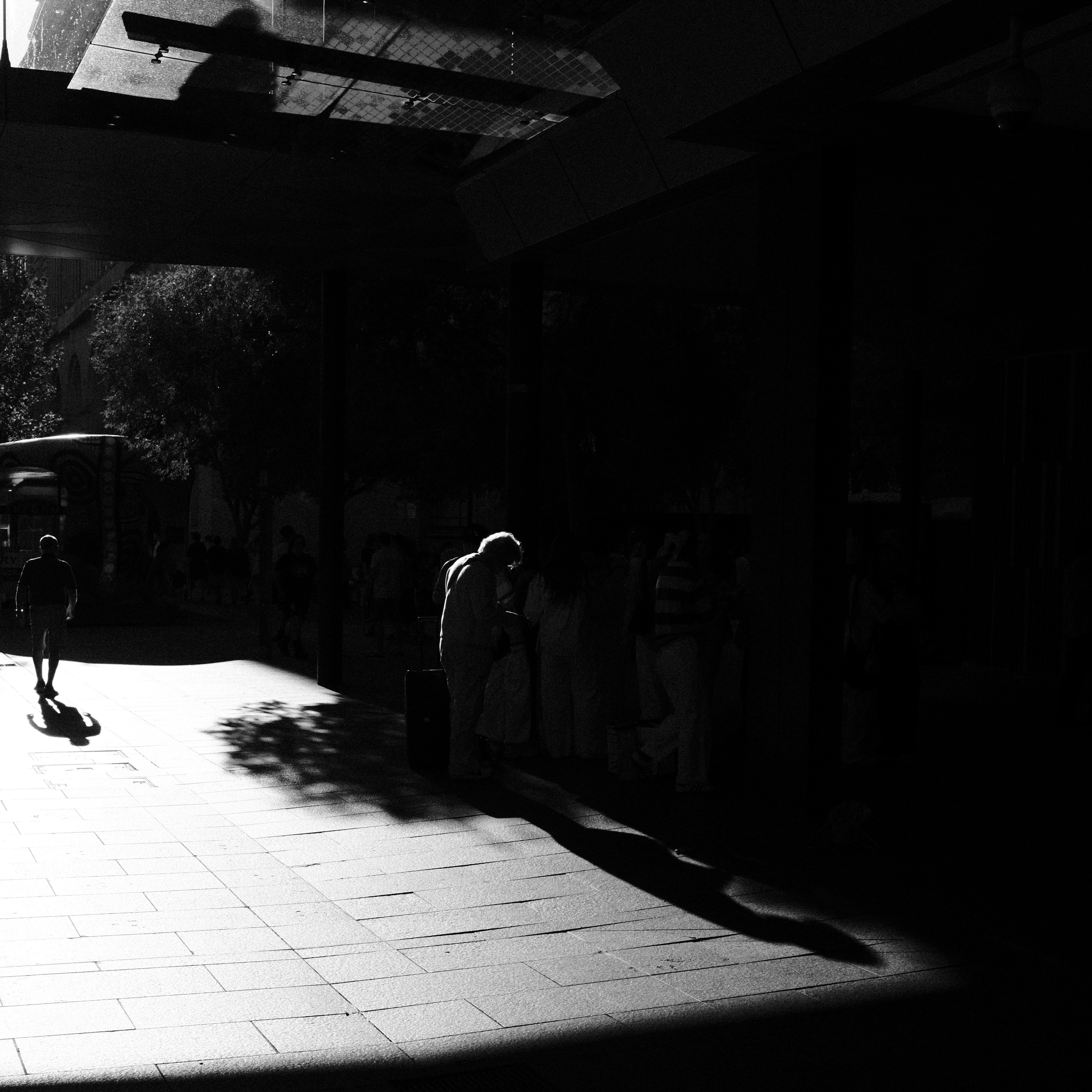 High-contrast black-and-white street scene under a building overhang, with bright sunlight on a tiled sidewalk. A lone pedestrian walks away on the left casting a long shadow, while several people stand in deep shade along the right wall; trees and a large vehicle are visible in the background.