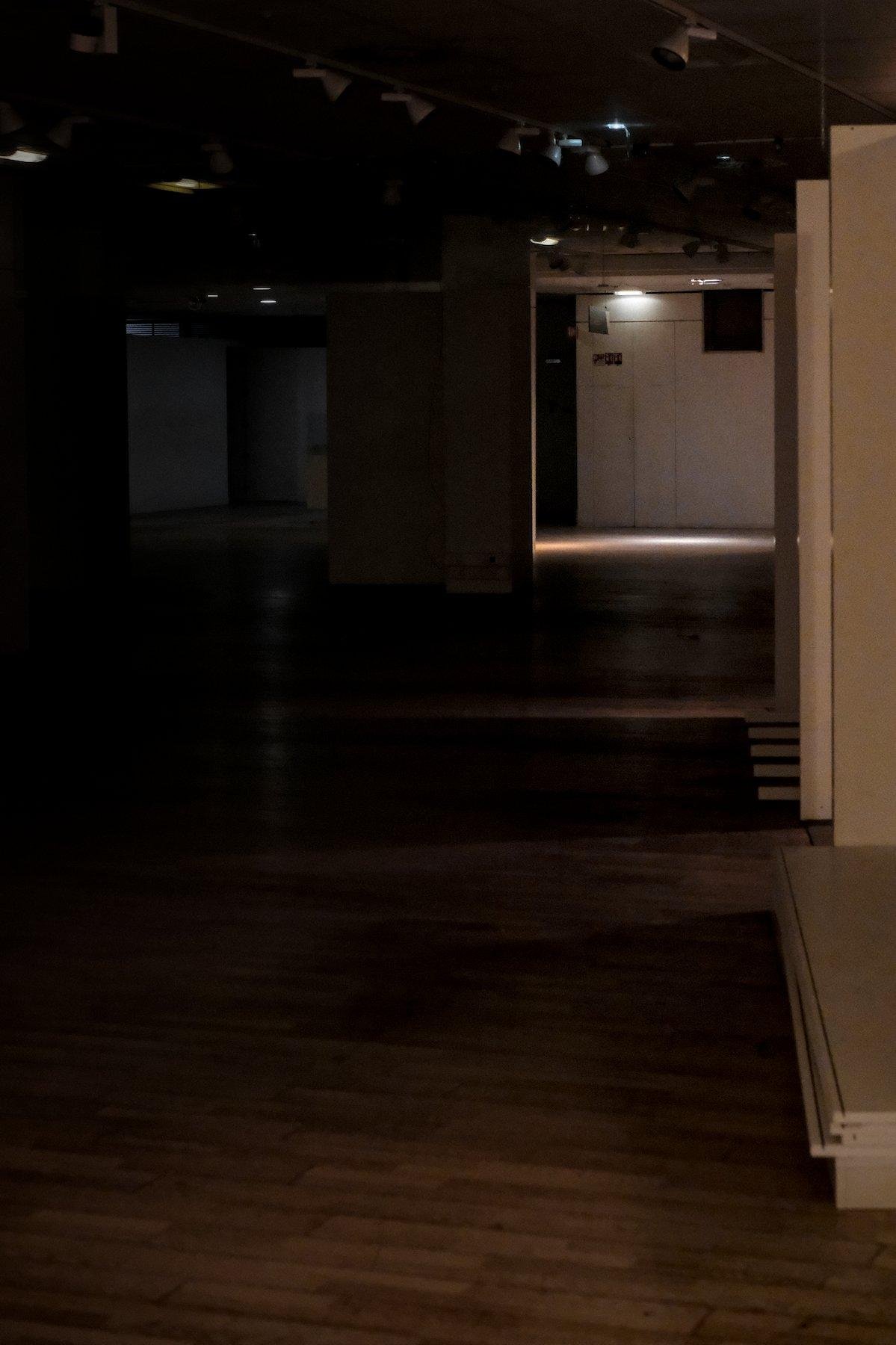 Photo verticale. Un endroit grand et sombre, au plancher vide. Sur la droite, des piliers blanc alignés, éclairés par une faible lumière orange, à gauche, la pénombre, au fond, une faible lampe éclaire le sol et le mur blanc qui semble avoir une porte cachée de service style armoire électrique