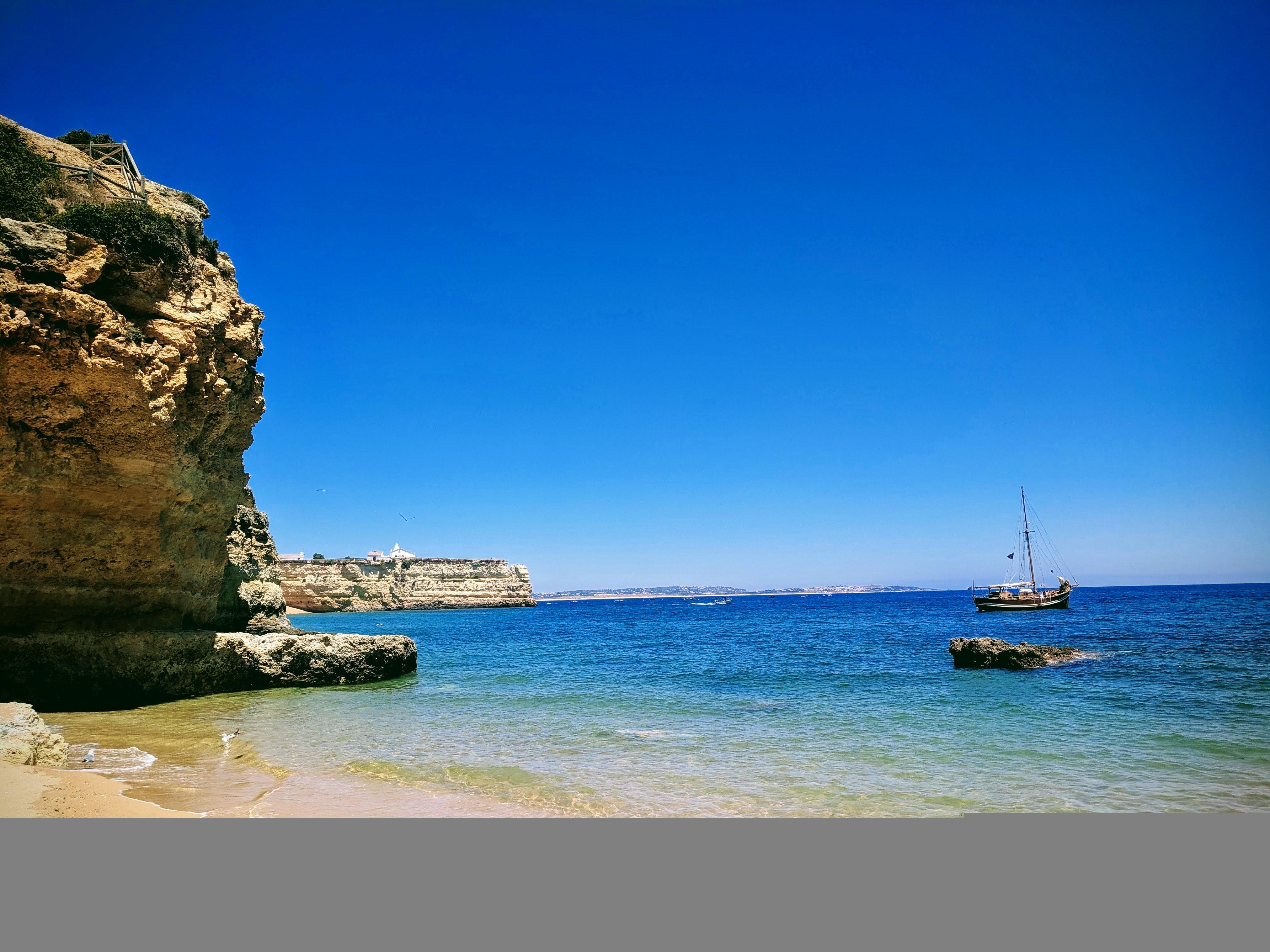 The image depicts a beautiful coastal scene. It features a rocky cliff on the left, a sandy beach with clear water, and a sailboat in the distance on the right. The sky is clear and blue, suggesting a sunny day.