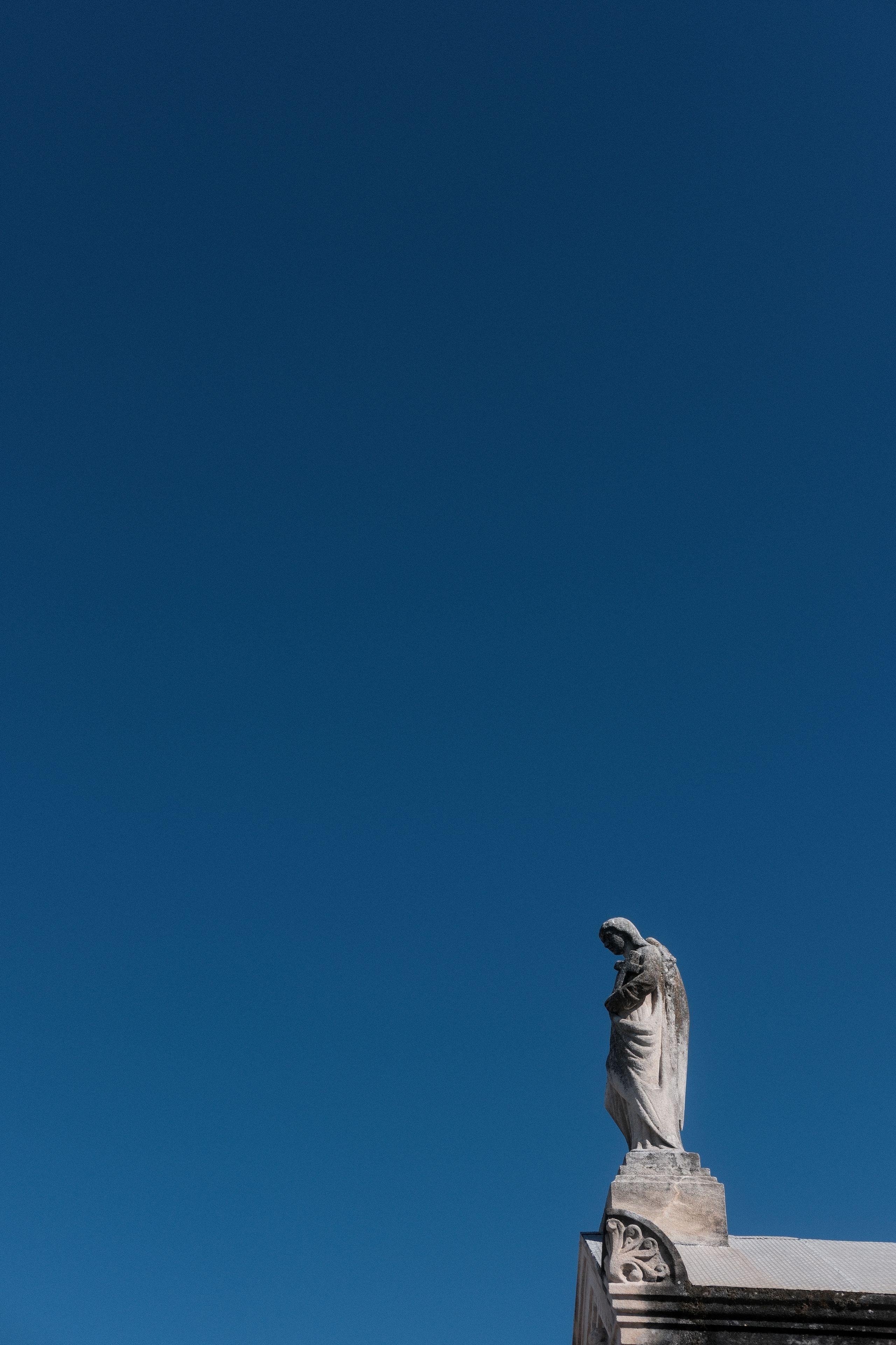 Photo verticale. L’image est majoritairement un ciel bleu avec dans l’angle inférieur droit, une statue blanche salies perchée en haut du toit d’un mausolée hors cadre