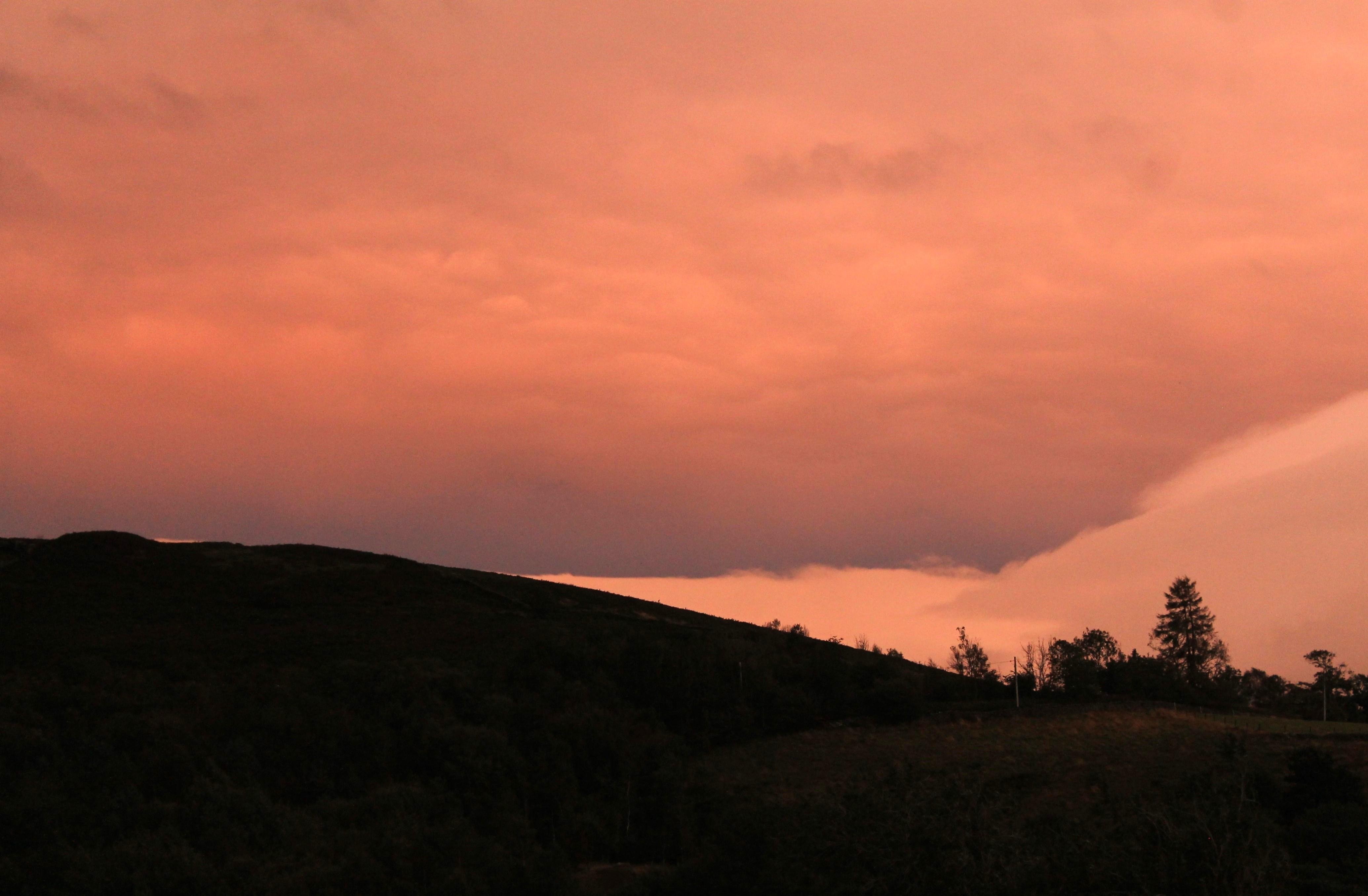 Una fotografía de un paisaje en pleno atardecer en el norte de Inglaterra. El cielo se ve naranja y repleto de nubes que generan texturas y capas . Se genera un contraste entre unas nubes más cercanas y más iluminadas que las del fondo, desde donde surge un degradado de oscuro a claro. El horizonte son colinas en una penumbra casi total, advertiéndose la silueta de unos árboles y una parte de hierba levemente iluminada