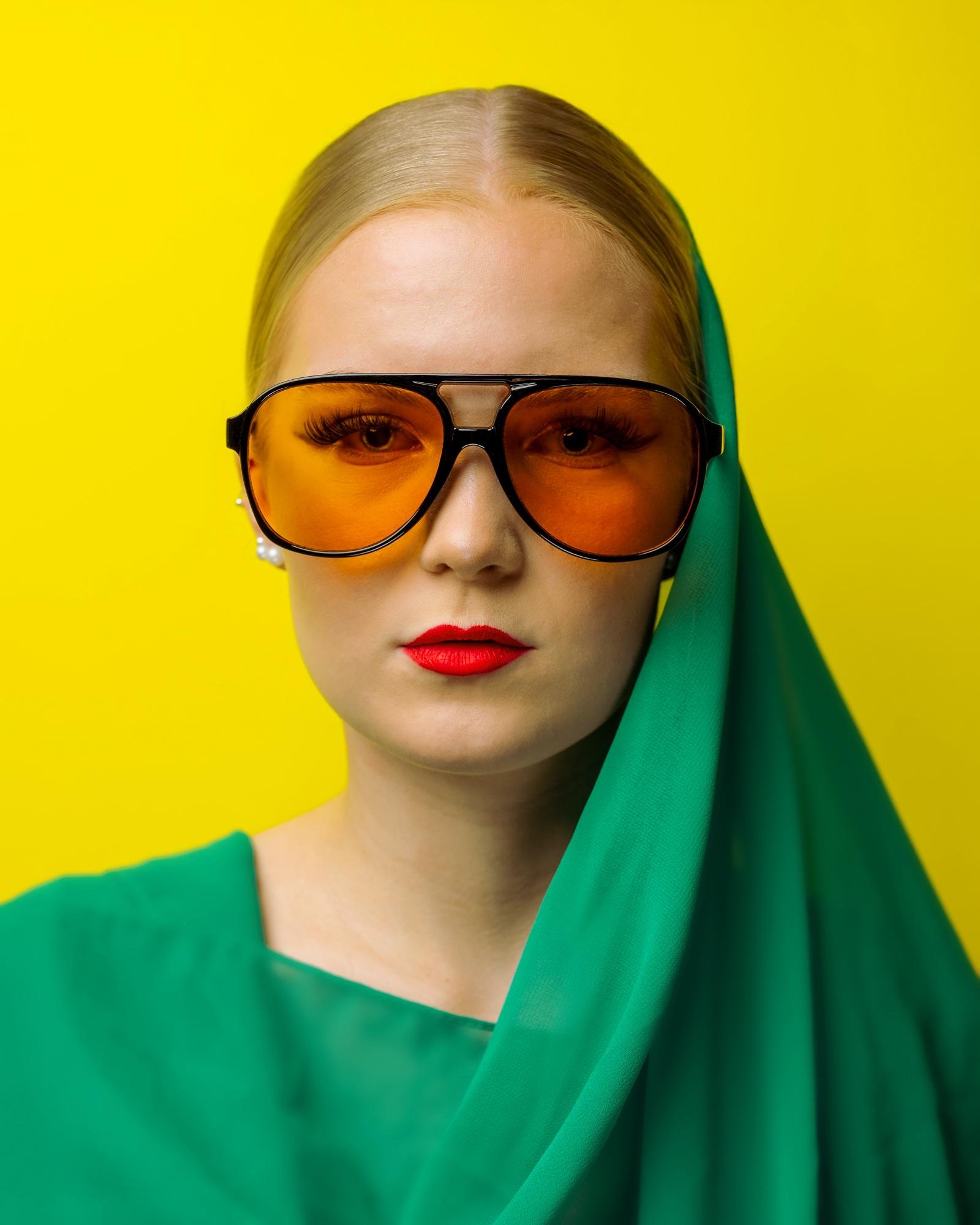 Centered close-up portrait of a blond person wearing oversized orange-tinted aviator sunglasses, red lipstick, and a green veil and top against a solid yellow background. Jarno.S Valokuvaaja Lappeenrannasta Kuvaajankulma