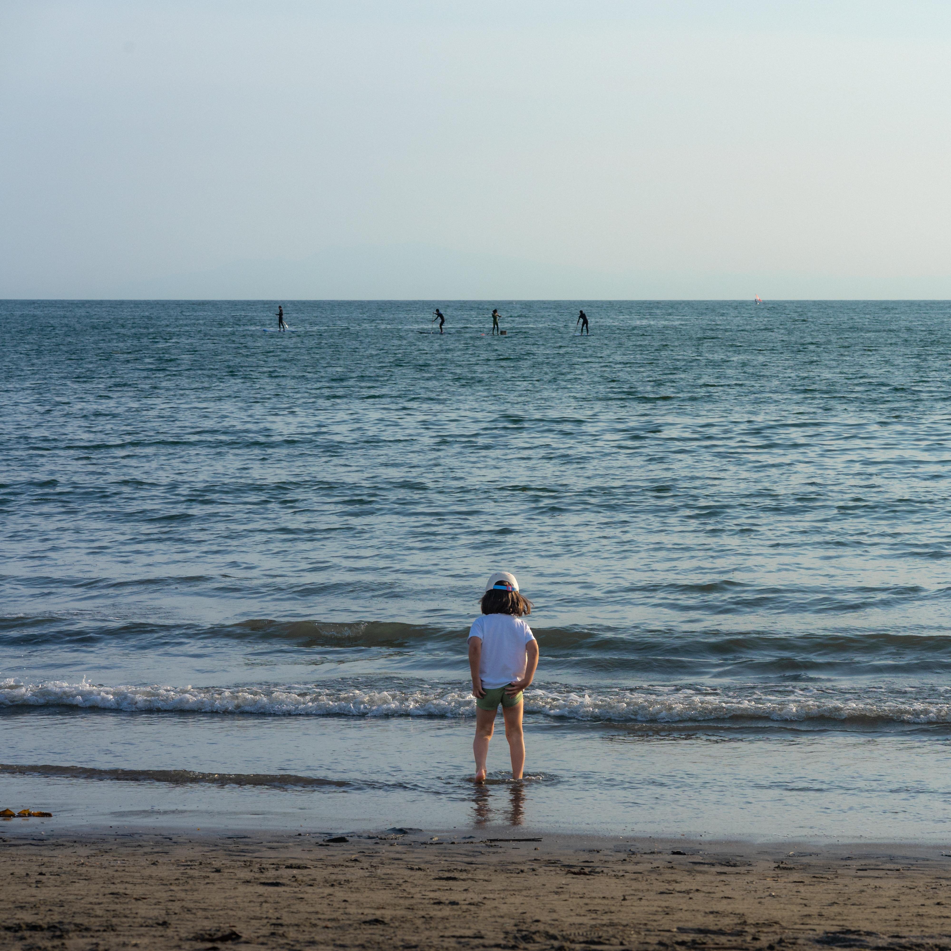 The image depicts a beach scene. There's a child standing near the shoreline, looking out at the water. In the background, several people are paddleboarding on the ocean. The weather appears to be clear and sunny.