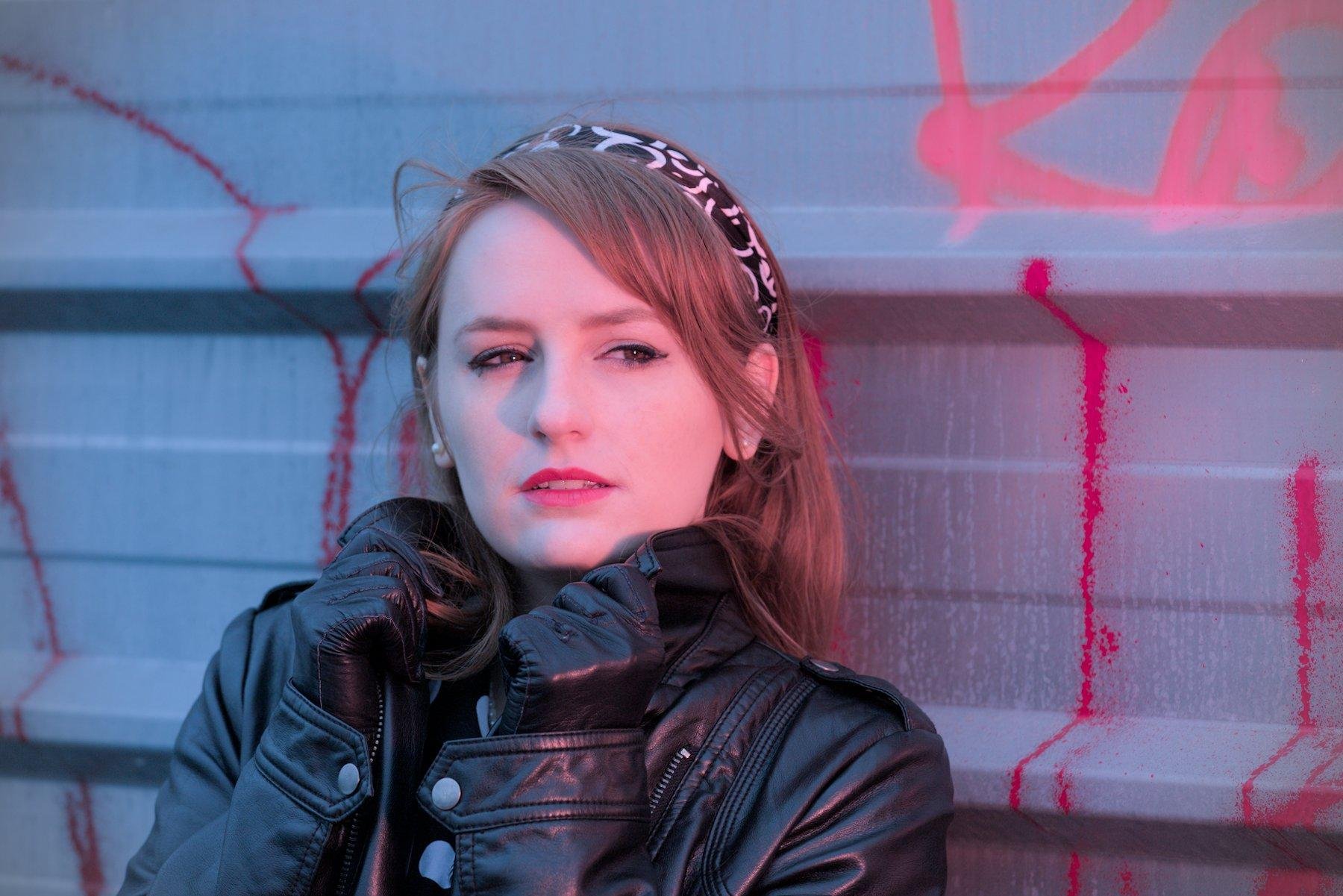 Photo format paysage d’une jeune femme rousse au cheveux long, coiffée d’un bandeau noir et blanc, portant un blouson noir en cuir, des gants noir en cuir, qui remonte le col du blouson sur son cou, en regardant légèrement sur sa gauche. La scène est rosée sur la droite de l’image et bleu sur la gauche. Elle est appuyée sur un mur en métal de chantier qui est taggé un peu partout