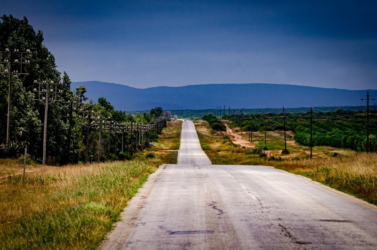 The image shows a long, straight road stretching into the distance. The road is flanked by grass on both sides, with utility poles running along the left side. In the background, there are hills or mountains under a cloudy sky. The scene has a rural and open feel.