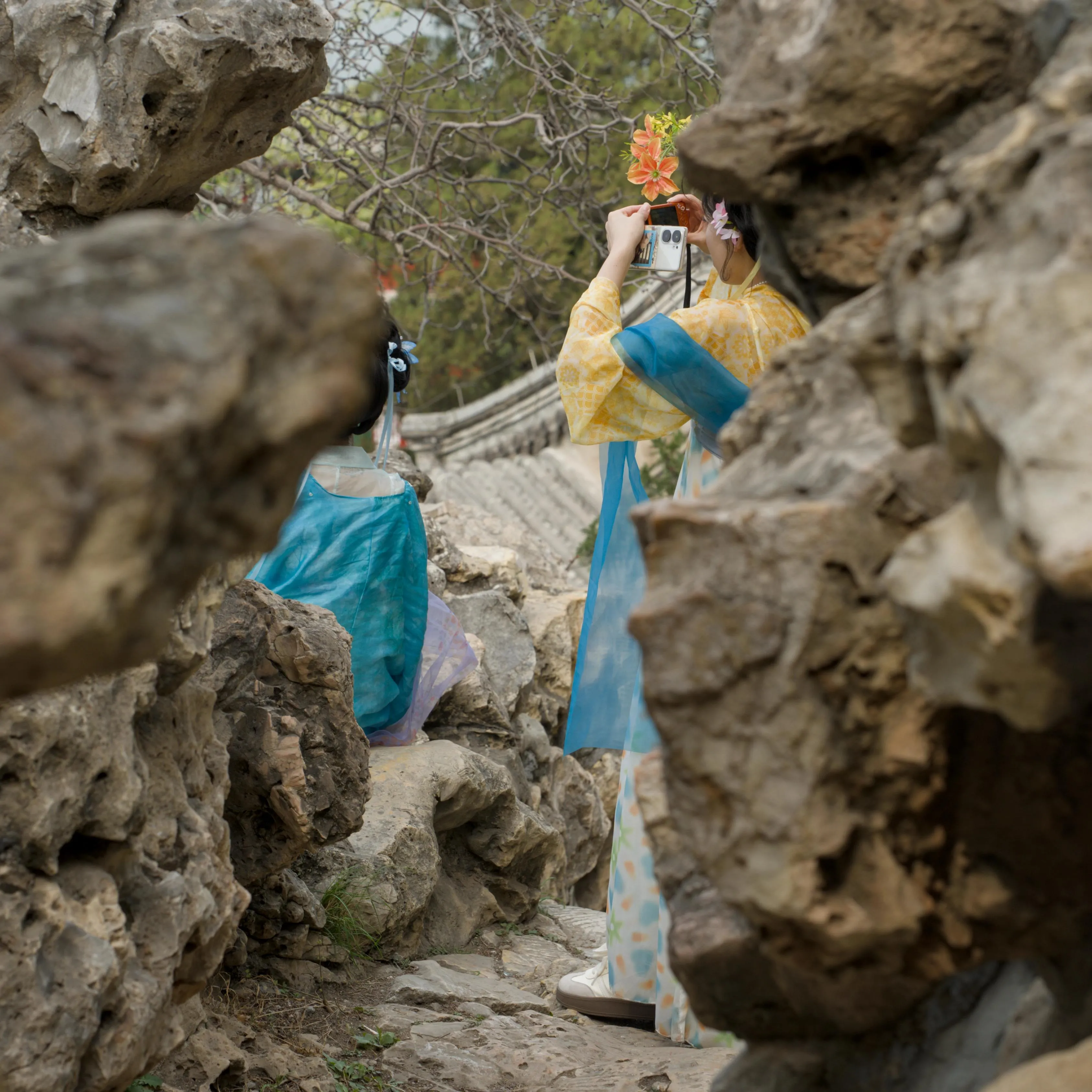 Photograph of two women wearing traditional-style Chinese long dresses, one sitting on a rock and posing and the second taking a photograph of the first using a compact digital camera, seen from their back. The dresses are yellow and blue. The women are on the other side of a path surrounded by a rocky wall. The rocks partially hide their features.