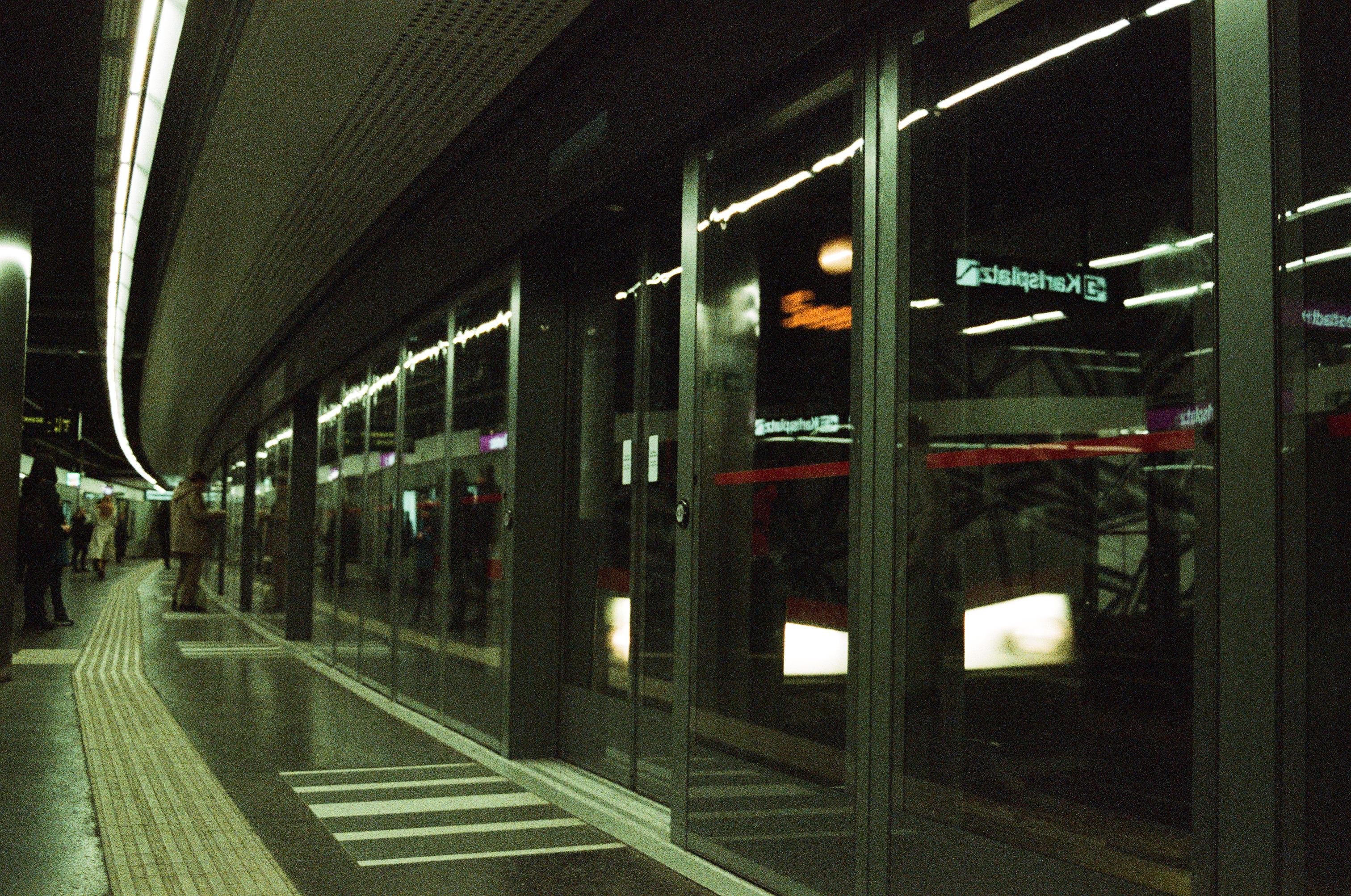 This image depicts a modern, indoor train or subway platform. There are glass doors separating the platform from the tracks, which reflects a common design for newer transit systems to enhance safety. The platform is mostly empty, with a few people visible in the distance. The lighting is artificial, typical for indoor transit stations.