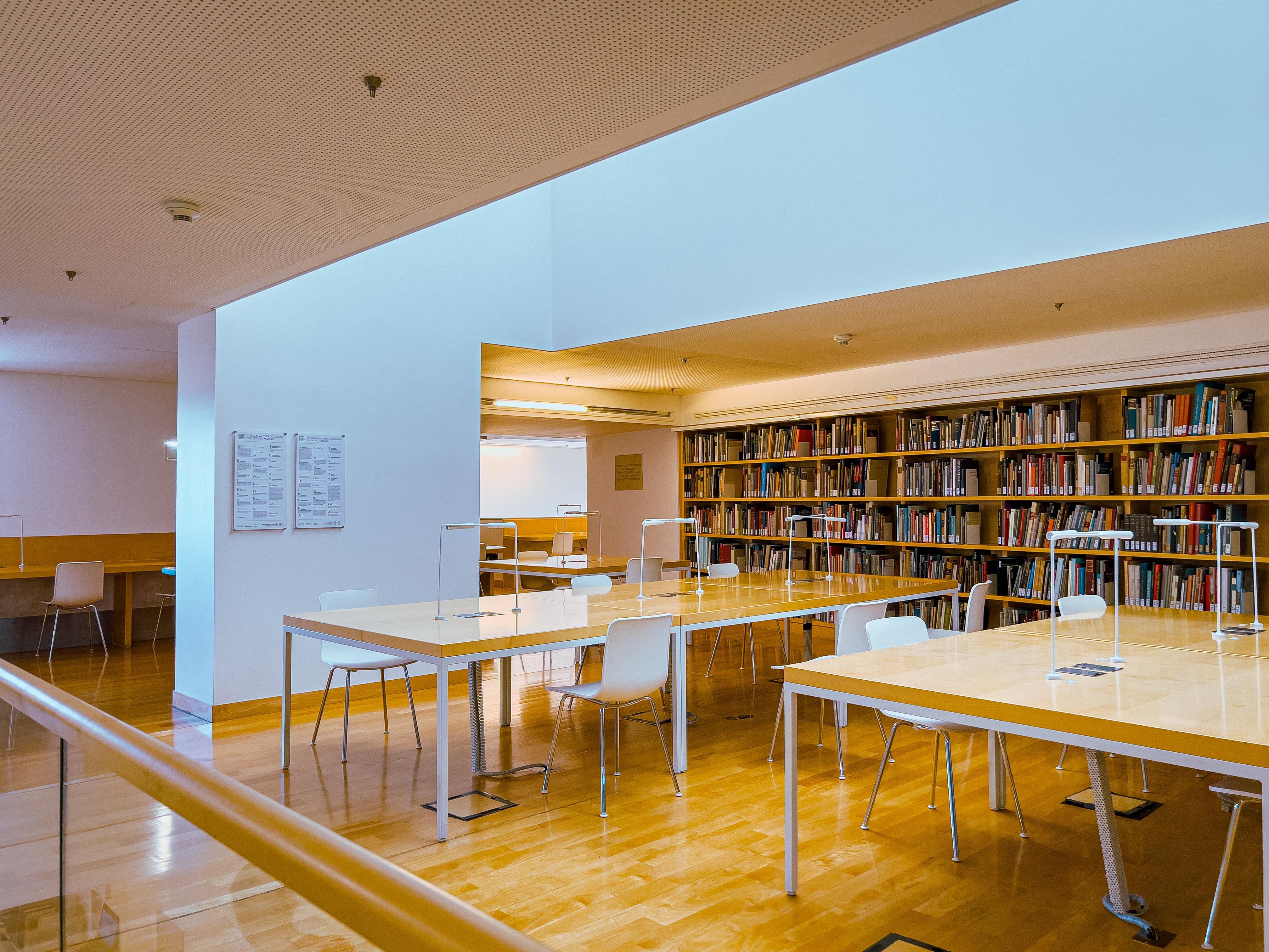 Una biblioteca o area di studio tranquilla. Ci sono diversi tavoli e sedie disposti per la lettura o lo studio, con scaffali pieni di libri lungo il muro. Lo spazio è ben illuminato, probabilmente dalla luce naturale che entra dal soffitto, ed è caratterizzato da un design minimalista con pavimenti in legno e pareti bianche.