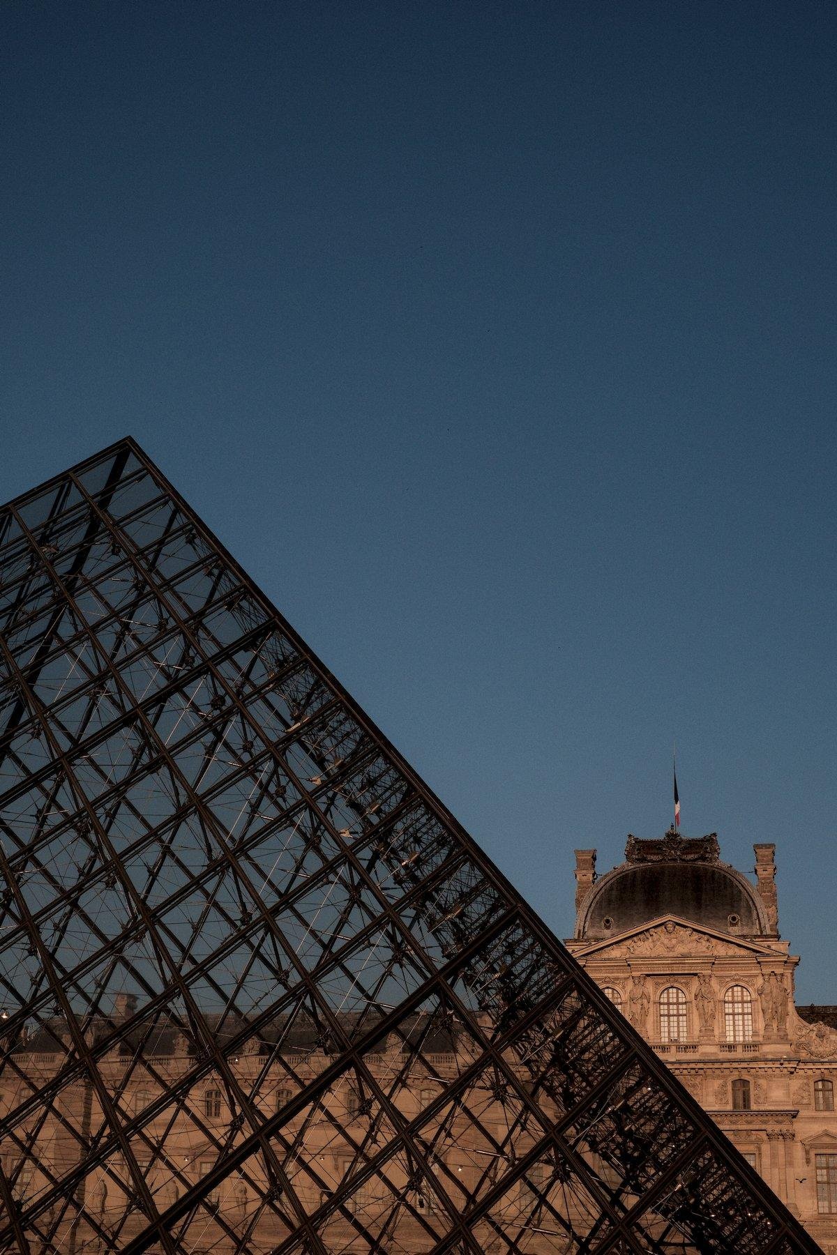 Photo verticale. Au premier plan, un bout de la pyramide du Louvre, avec le Louvre derrière éclairé par le soleil couchant le rendant orangé, sous un ciel en dégradé de bleu