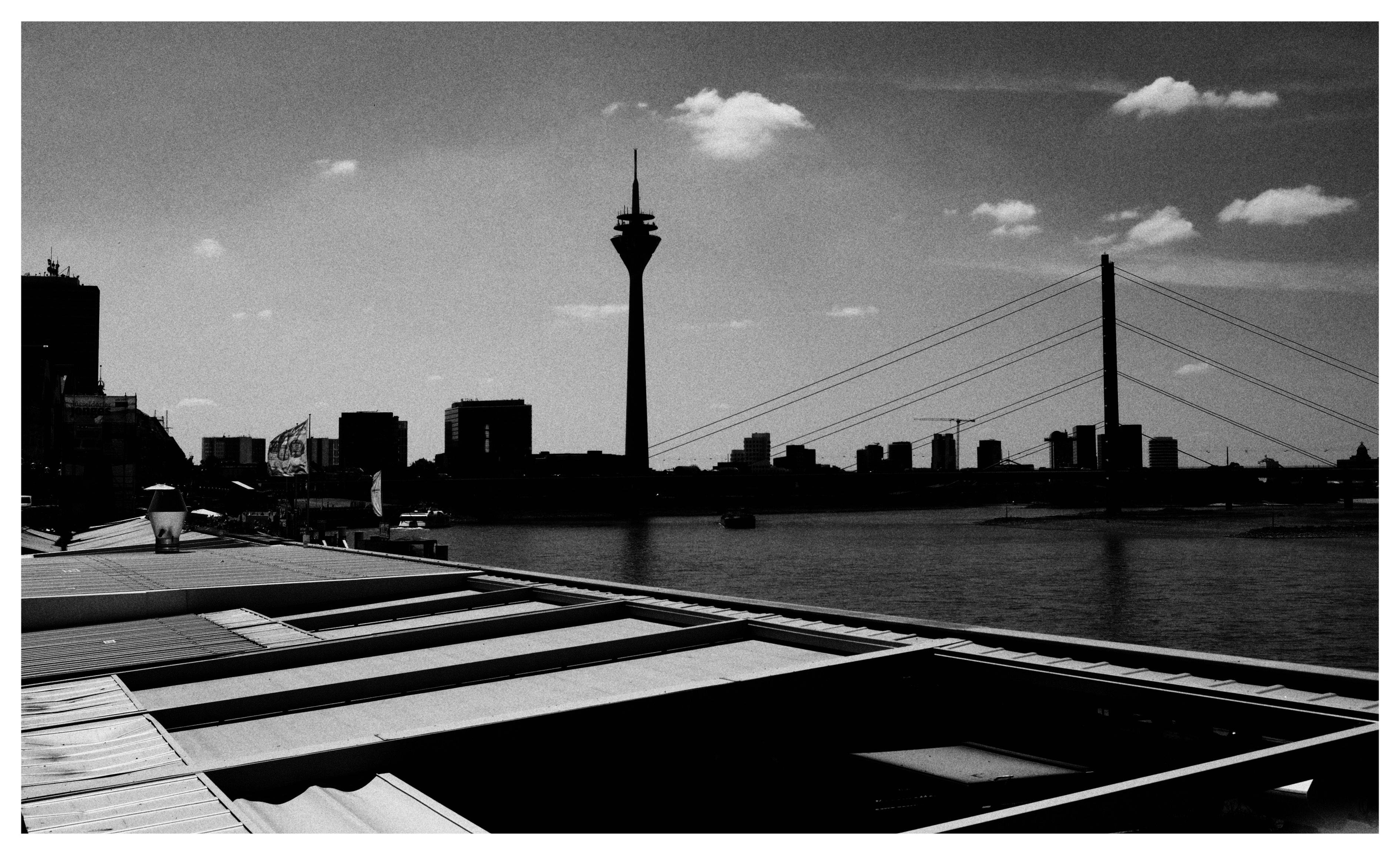 Black-and-white waterfront city skyline with a tall observation tower near center and a cable-stayed bridge to the right, seen across a river; angular roof structures and a walkway with small flags in the foreground under a partly cloudy sky.