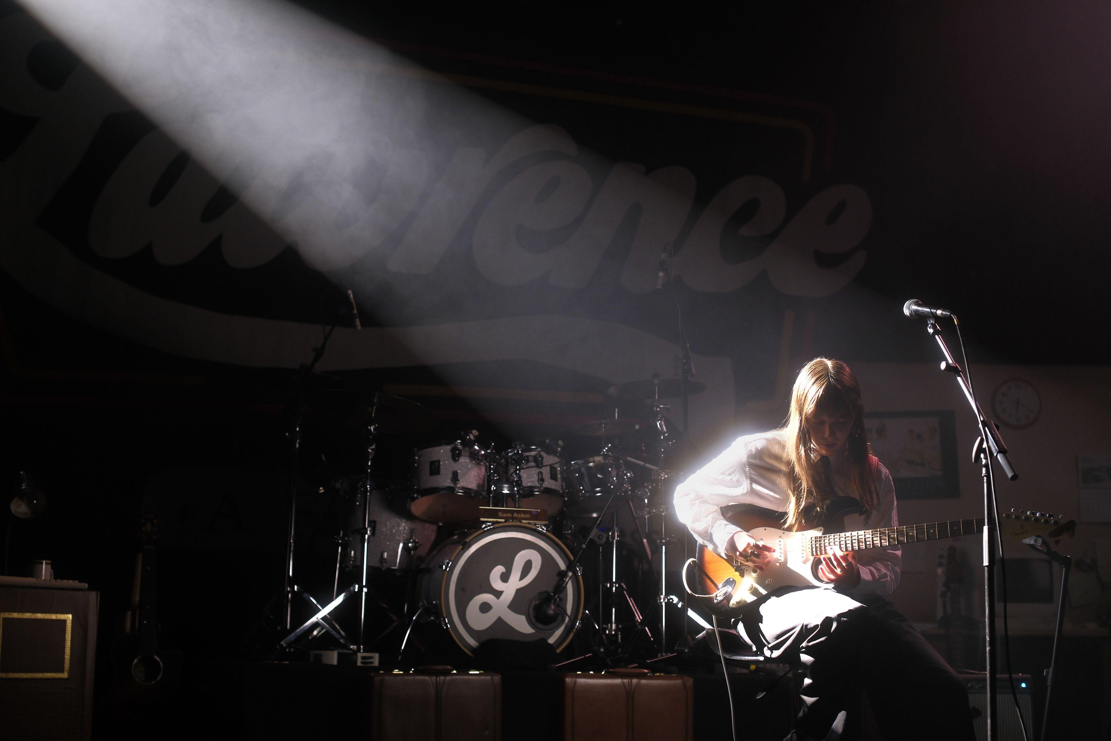 Photo format paysage montrant une musicienne au cheveux long, assise, concentrée sur sa guitare électrique, portant une chemise blanche et un pantalon noir, éclairée par un spot blanc