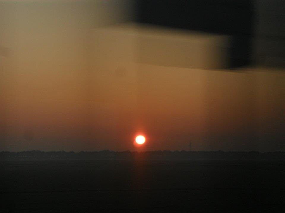Una foto hecha desde un tren en movimiento, de una puesta de sol oscura y anaranjada, con un sol grande, brillante y naranja casi rozando el horizonte. Se ven siluetas en movimiento de señales de tráfico