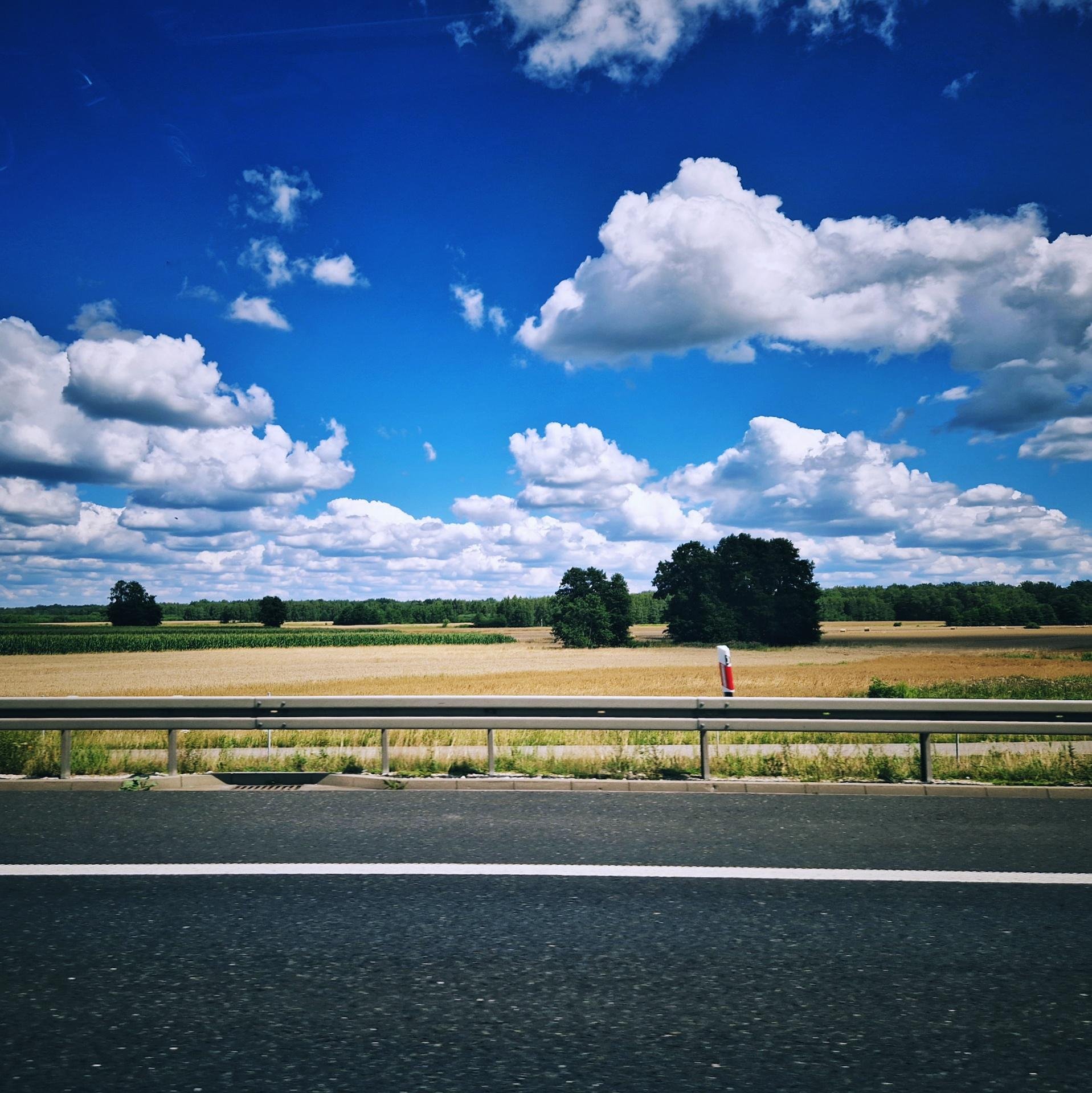 Open road with a metal guardrail in the foreground, dividing a field. Greenery and trees are visible in the middle ground. Bright blue sky is filled with scattered fluffy white clouds.