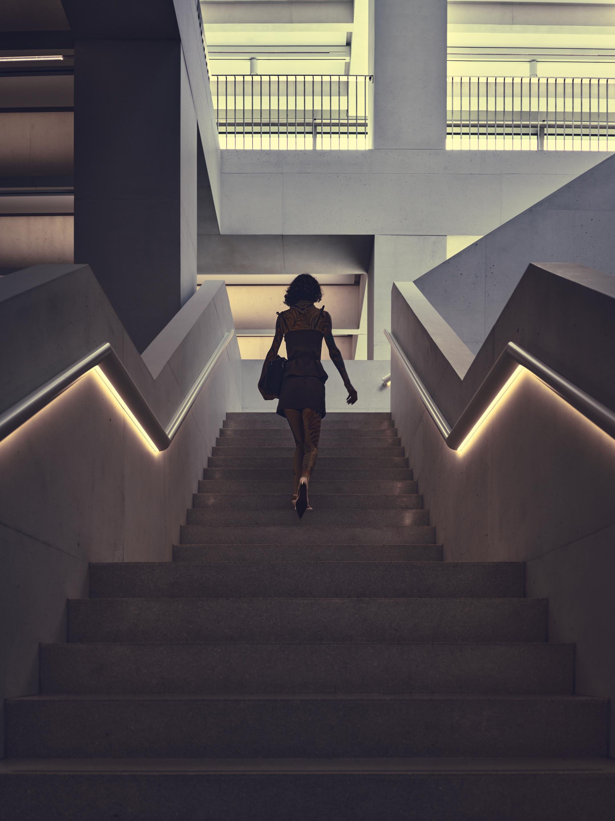 Person walking up a wide concrete staircase in a modern building, centered in the frame, with illuminated handrails on both sides and geometric upper-level railings and beams overhead.