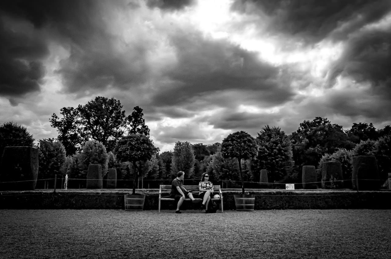 Two people sit on a bench facing each other in a landscaped park. The surrounding area is lined with trees and shrubs. Overhead, dramatic, dark clouds fill the sky, creating a stark contrast with the lighter ground.