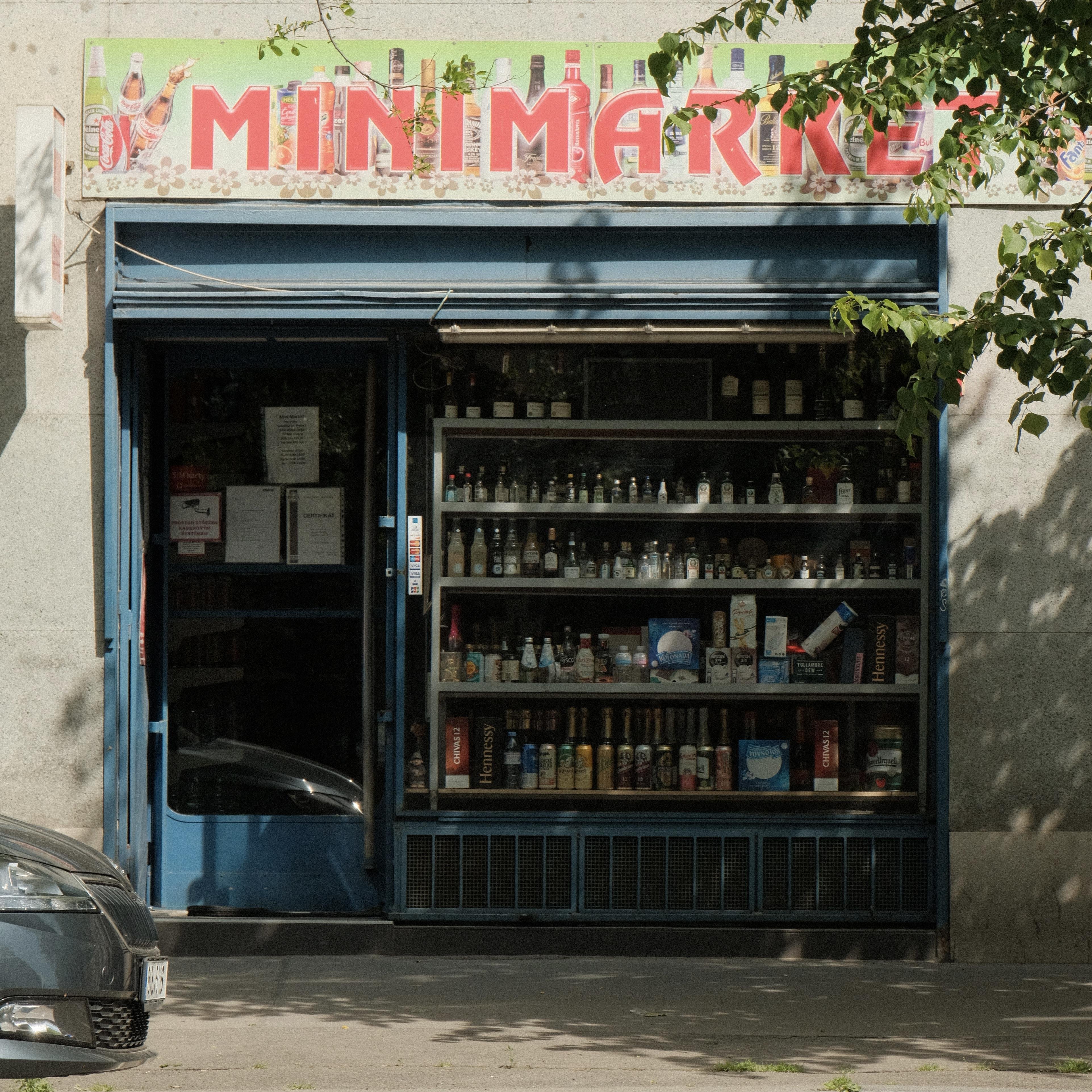 A square shop window of a mini supermarket filled from top to bottom with bottles on shelves advertising many different drinks sold in the store.
To the left is the entrance door, above the window large red letters spell „MINIMARKET“.
