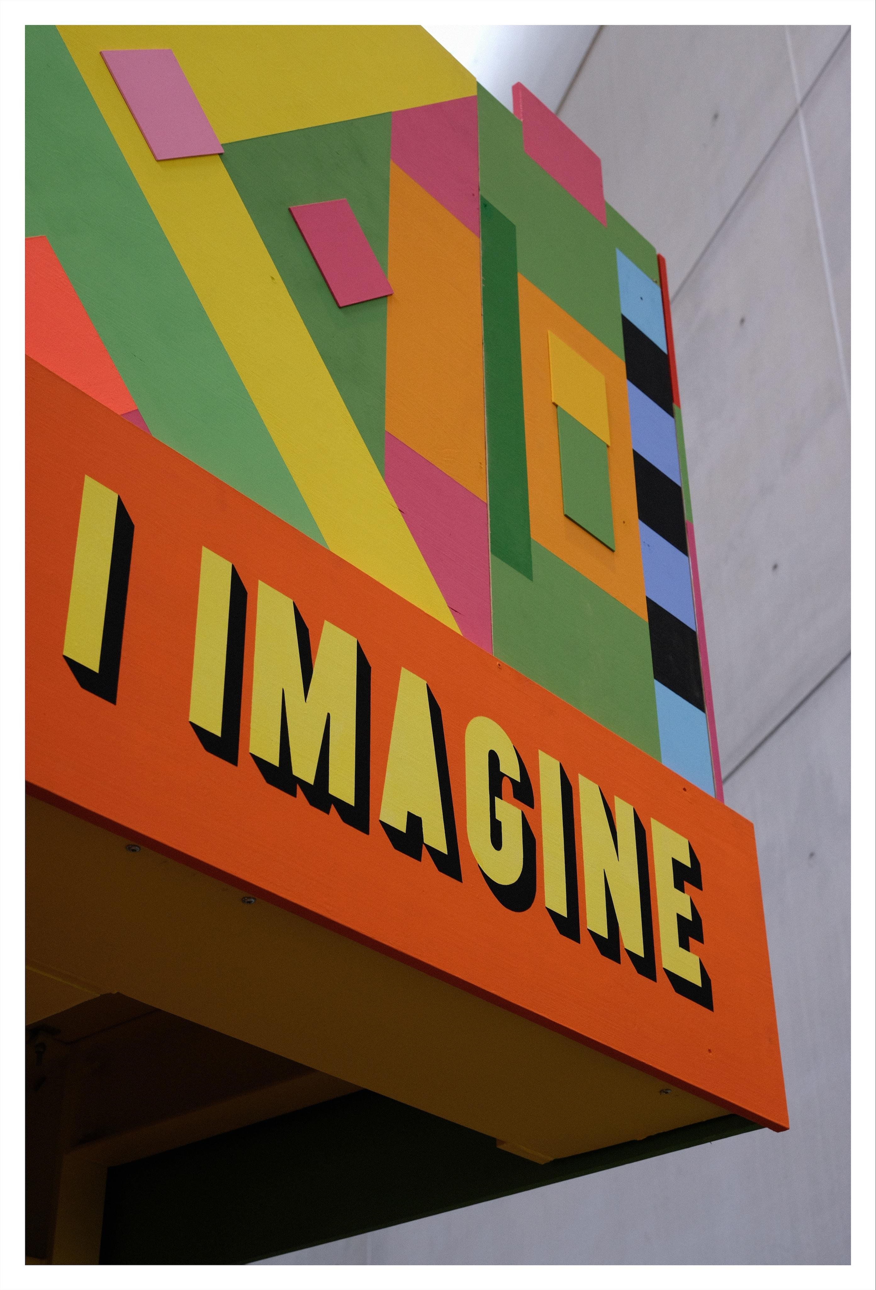 Low angle view of an indoor wooden balcony painted in different colors. In yellow large letters „I imagine“ is written across the balcony. A concrete wall in the background.