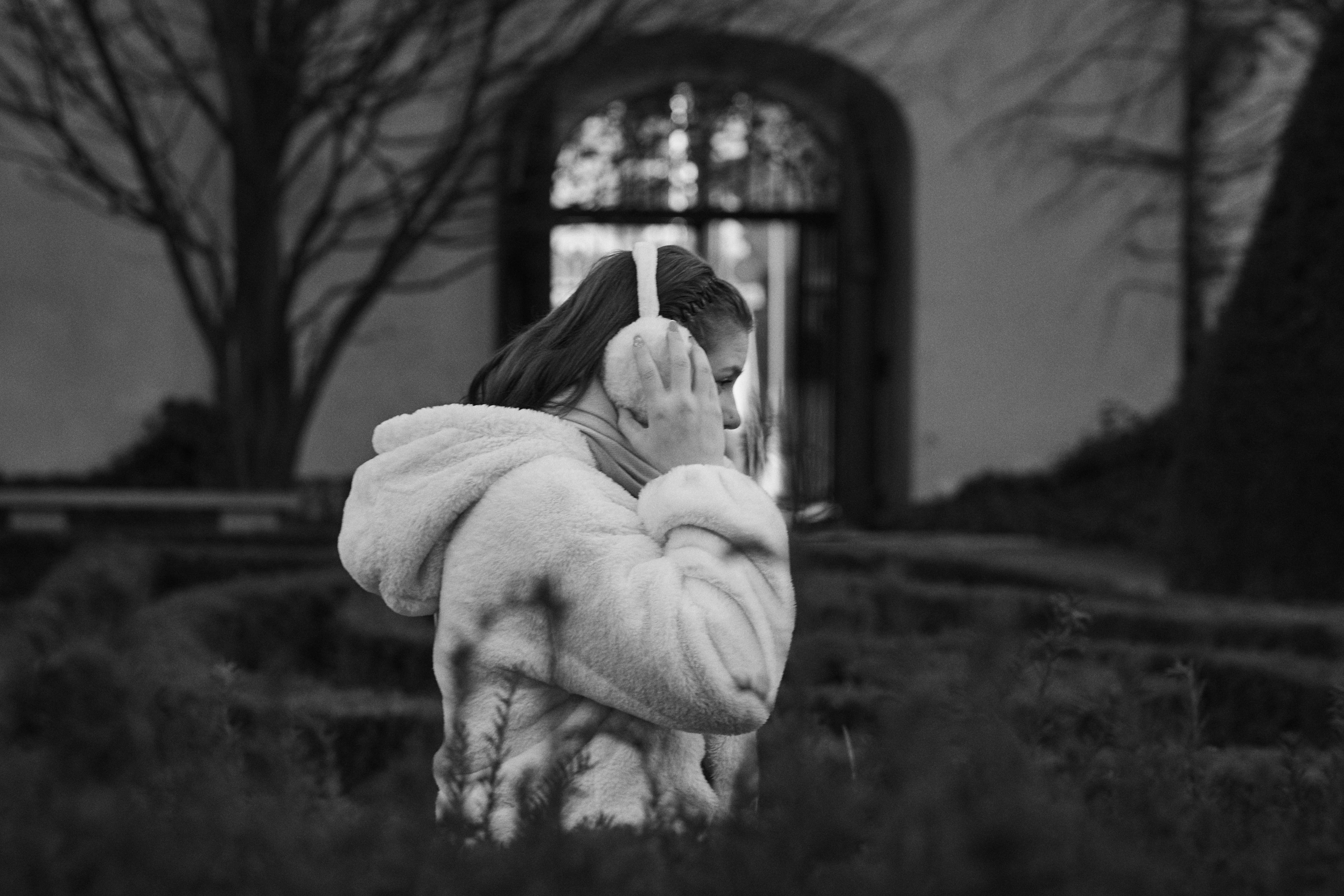 The image shows a person wearing a fluffy coat and earmuffs, walking in an outdoor setting. The background includes a garden or park-like area with trees and a prominent gate or arch. The scene is in black and white.