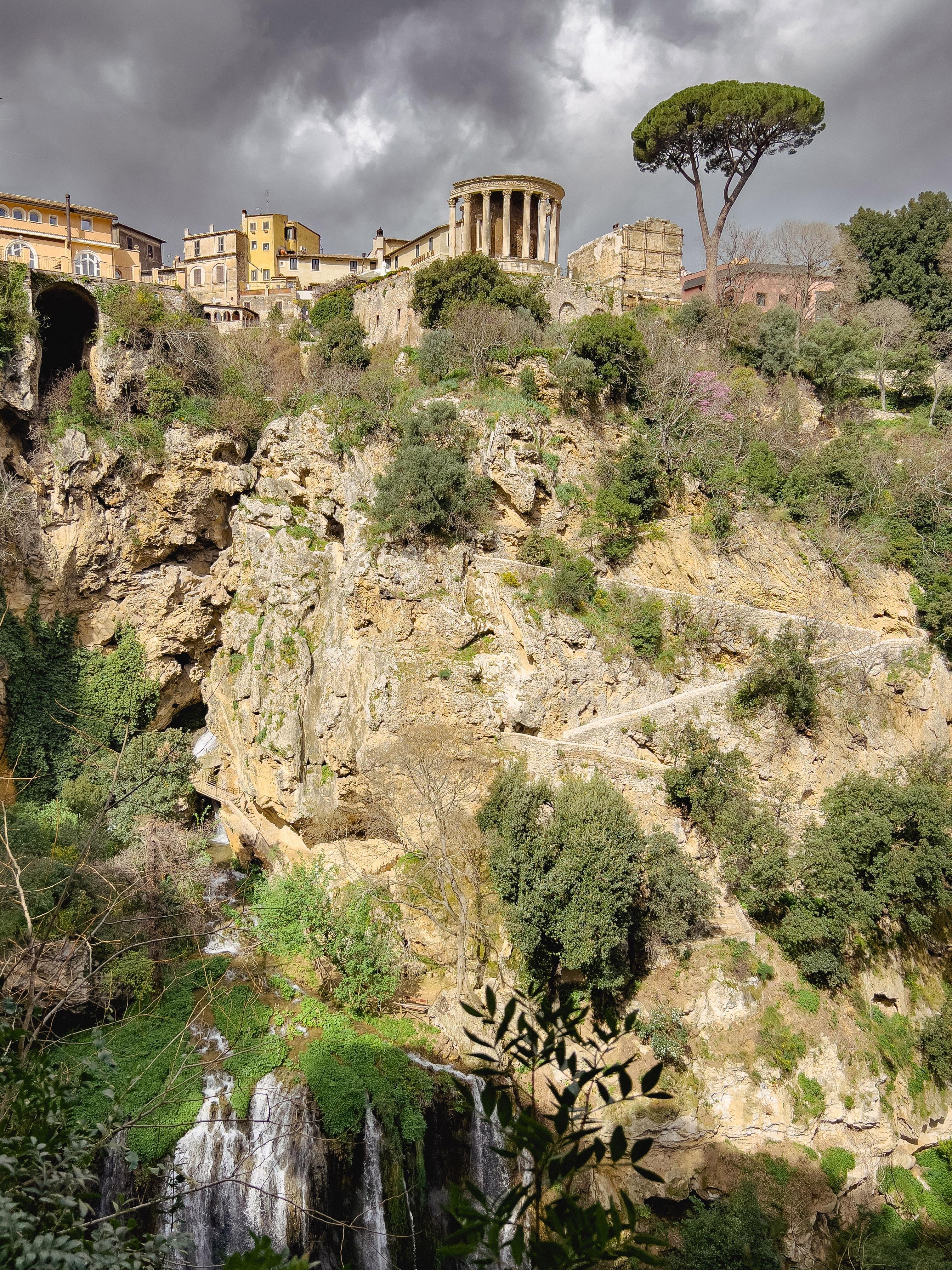 Paesaggio pittoresco con antiche strutture situate su un pendio roccioso. Prominente nell'immagine c'è un tempio circolare con colonne, noto come Tempio di Vesta, situato a Tivoli, in Italia. Intorno al tempio ci sono edifici in pietra e una rigogliosa vegetazione. Un cielo drammatico con nuvole scure aggiunge atmosfera alla scena e, nella parte inferiore dell'immagine, una cascata si precipita giù dalle rocce.