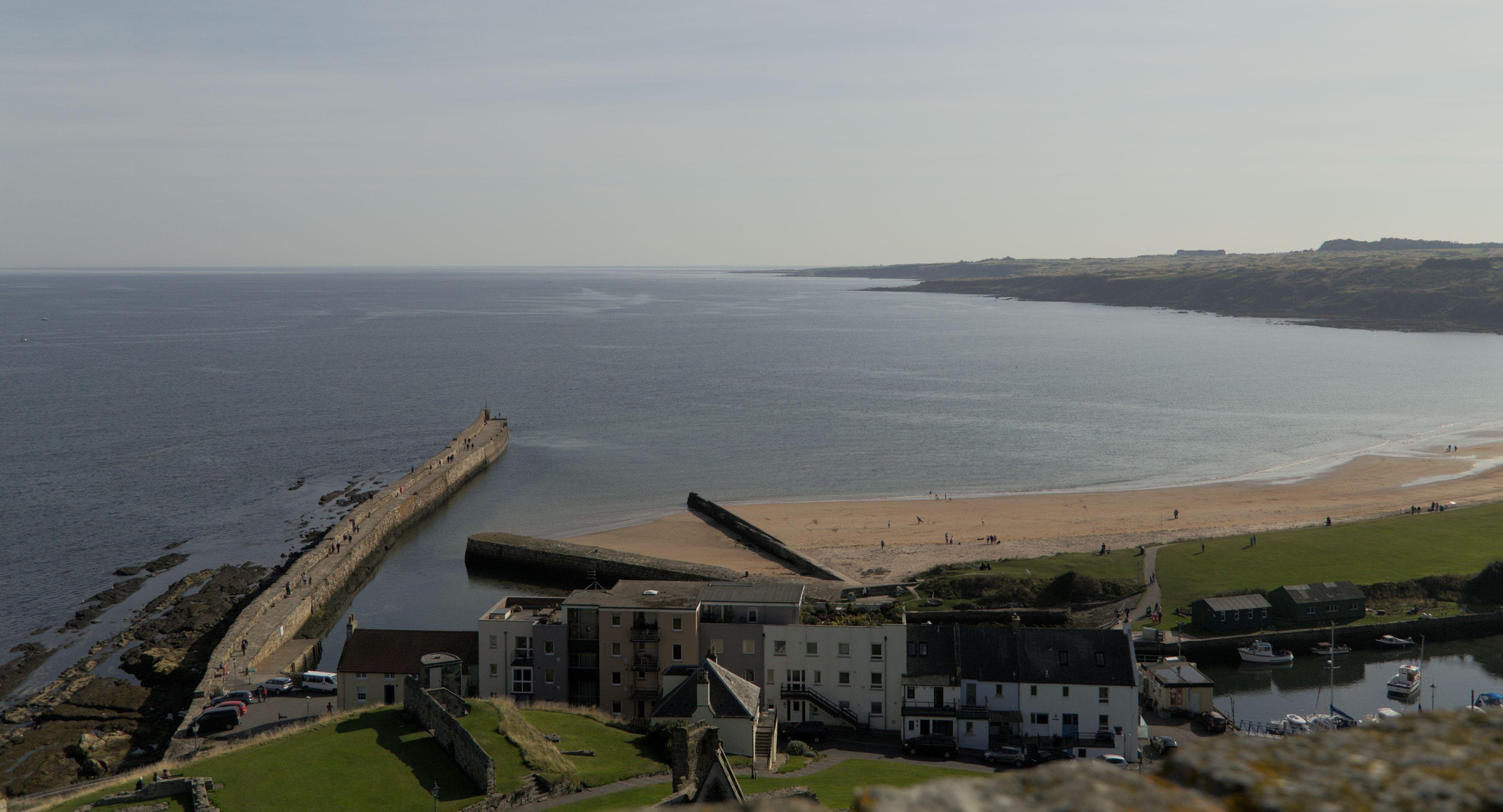 A photo from a high point of view of the city of St Andrews, Scotland. We can see the sea and an old now unused dock with tourists on it of an harbor, a sand beach and some grass. A piece of land is peaking in from the right, and the bottom is occupied by some buildings, The sky is very pale blue with a lot of clouds