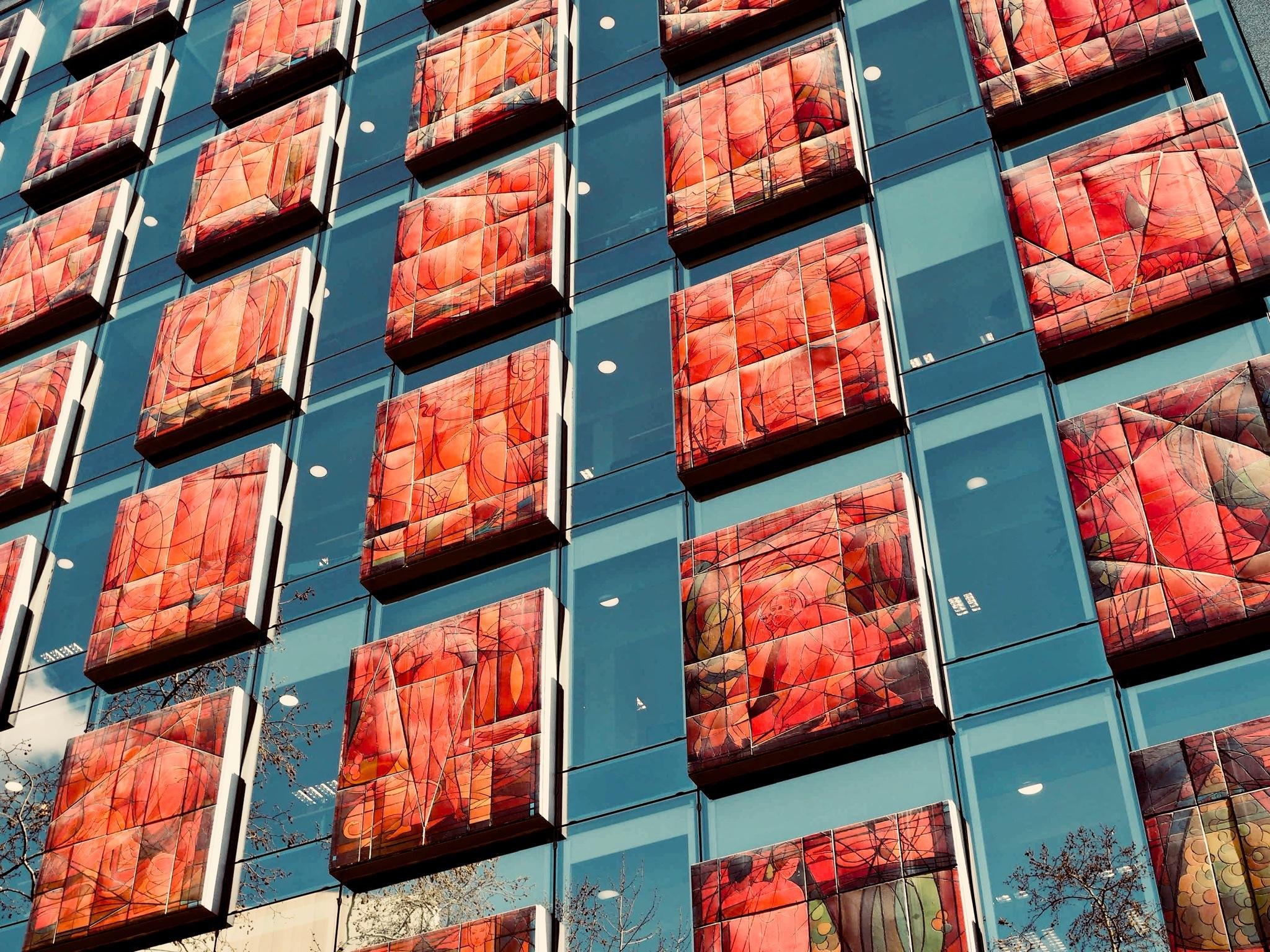 Abgewinkelte Ansicht einer modernen Glasgebäudefassade mit Reihen von hervorstehenden rechteckigen Paneelen, die jeweils rote und orangefarbene abstrakte Kunstwerke zeigen, getrennt durch blau getönte Fensterabschnitte mit kleinen weißen kreisförmigen Lichtern, die sich im Glas spiegeln.