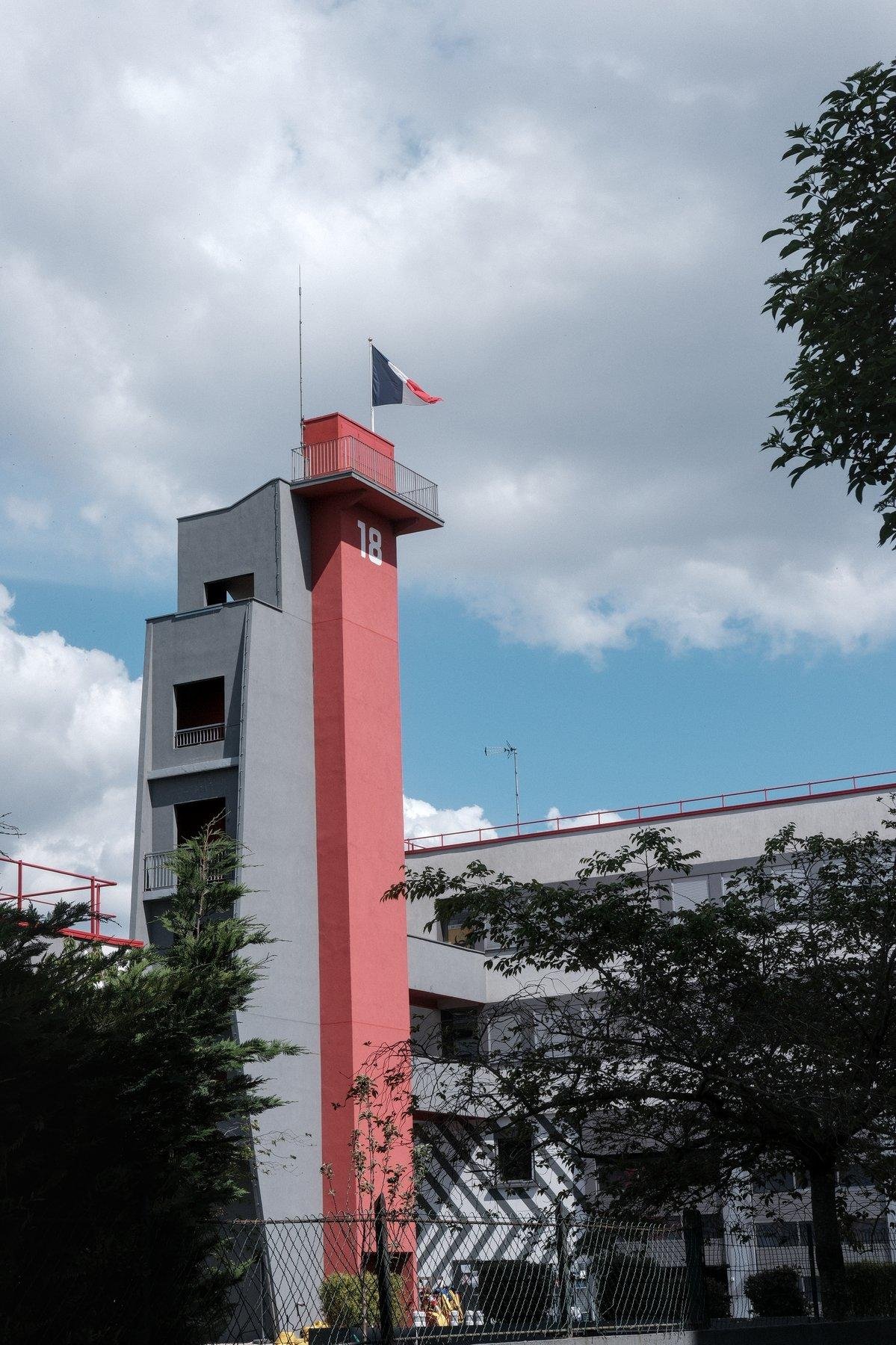 Photo verticale. Tour moderne grise et rouge, avec un drapeau français flottant au sommet et un gros « 18 » peint presque tout en haut, juste en dessous d’un poste d’observation